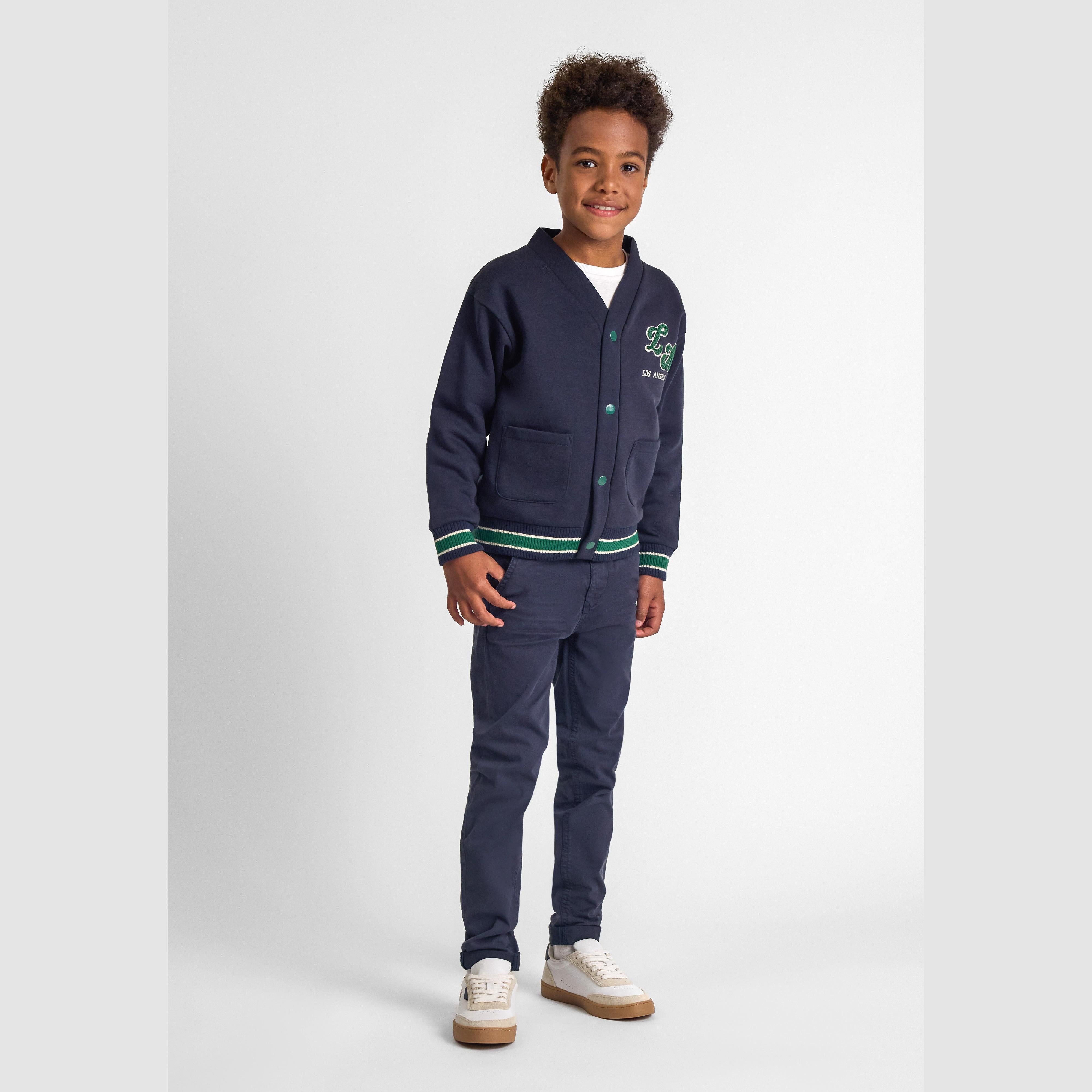 Young boy wearing navy cardigan with LA applique and matching trousers, standing against white background, full body front view