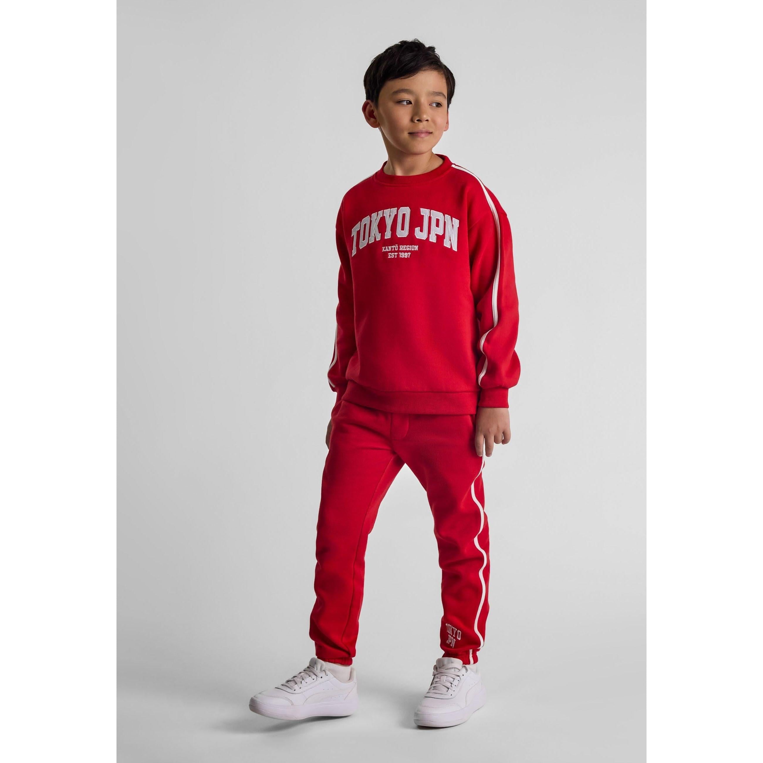 Young boy wearing red tracksuit set with Tokyo JPN embroidery, standing against grey background in white trainers