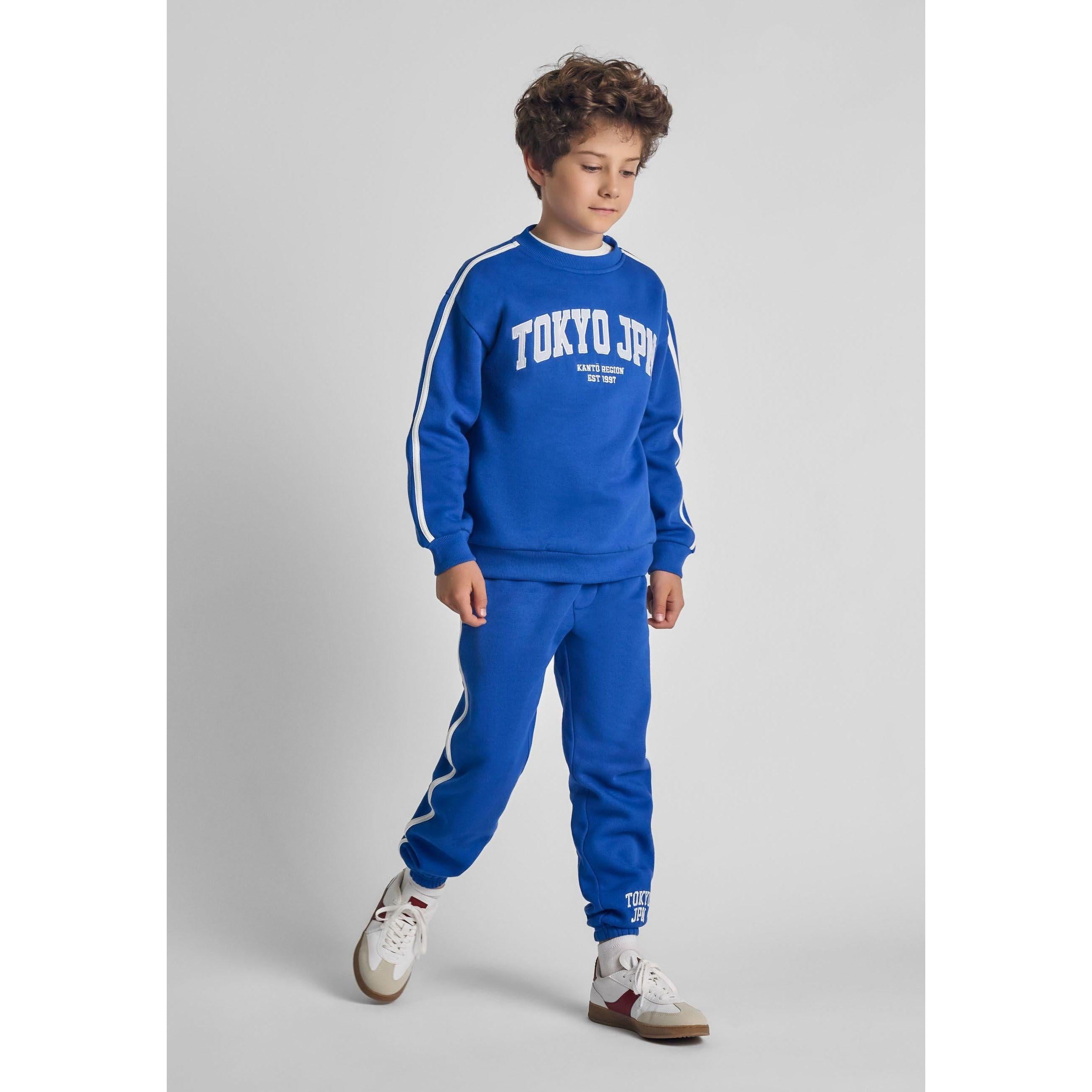Boy wearing cobalt blue tracksuit set with Tokyo JPN embroidery, white stripe details on sleeves, standing against grey background wearing white trainers