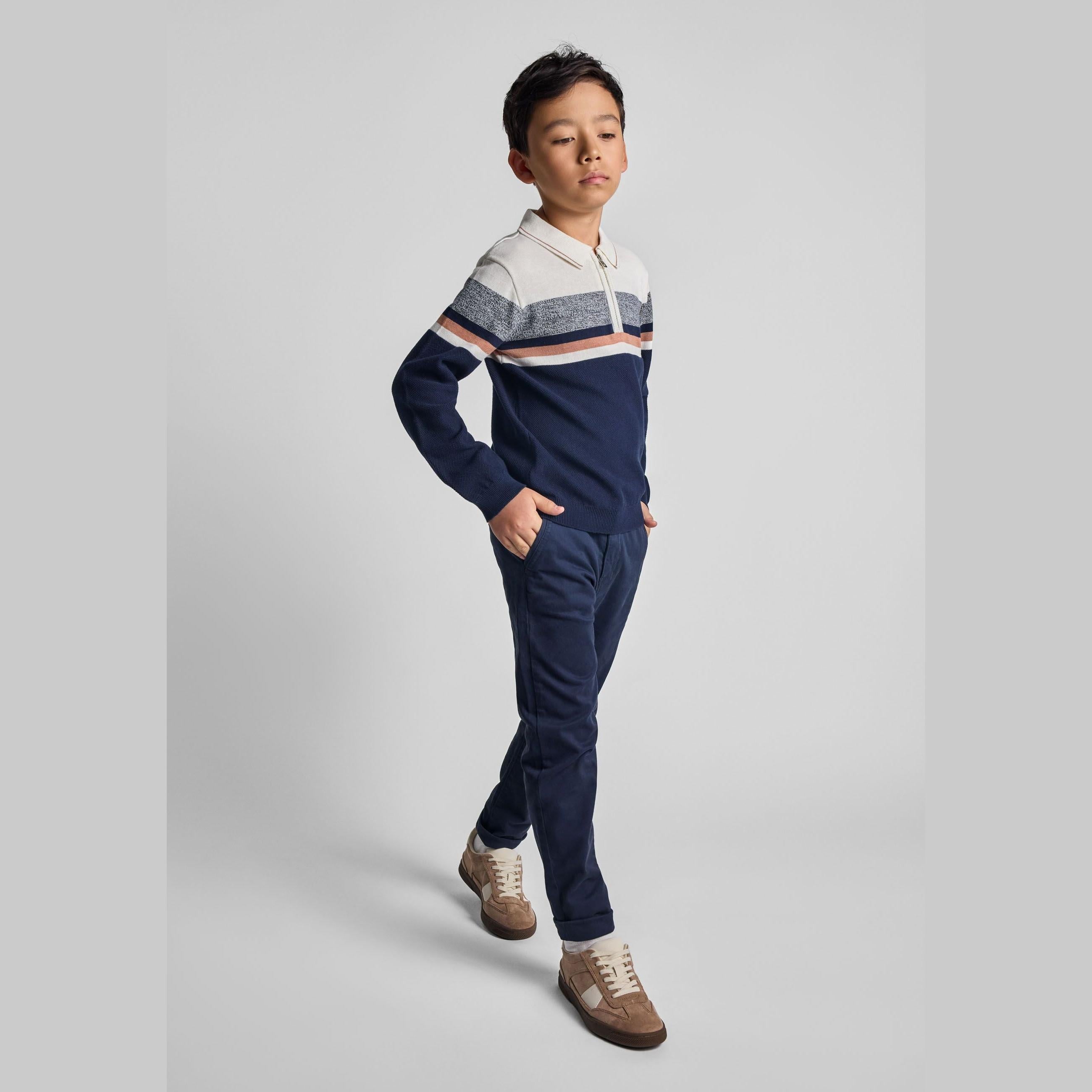 Young boy wearing cream and navy striped polo jumper with quarter zip and matching navy trousers, posed against light grey studio background