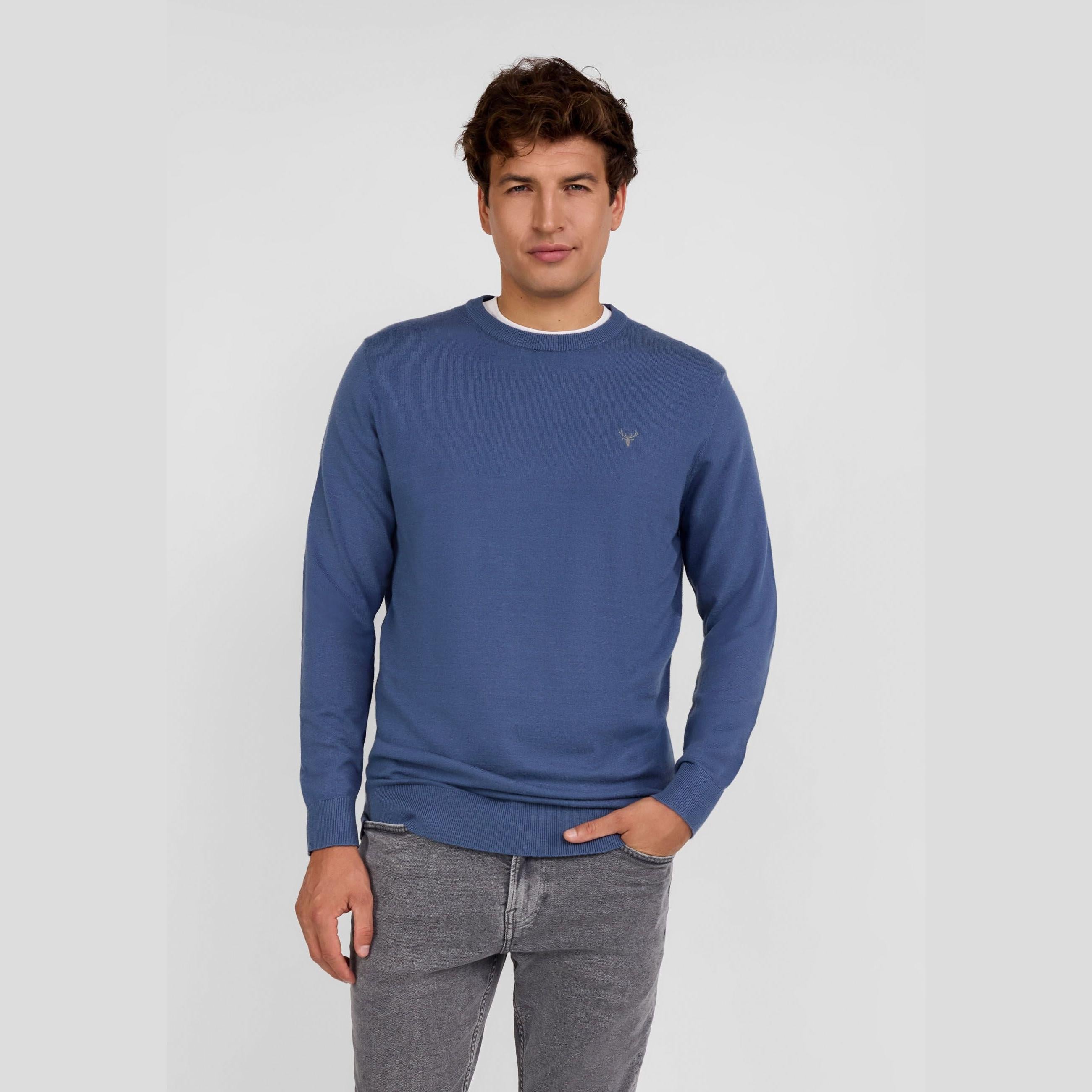 Man wearing blue fine knit crew neck jumper with grey jeans, front view against white background