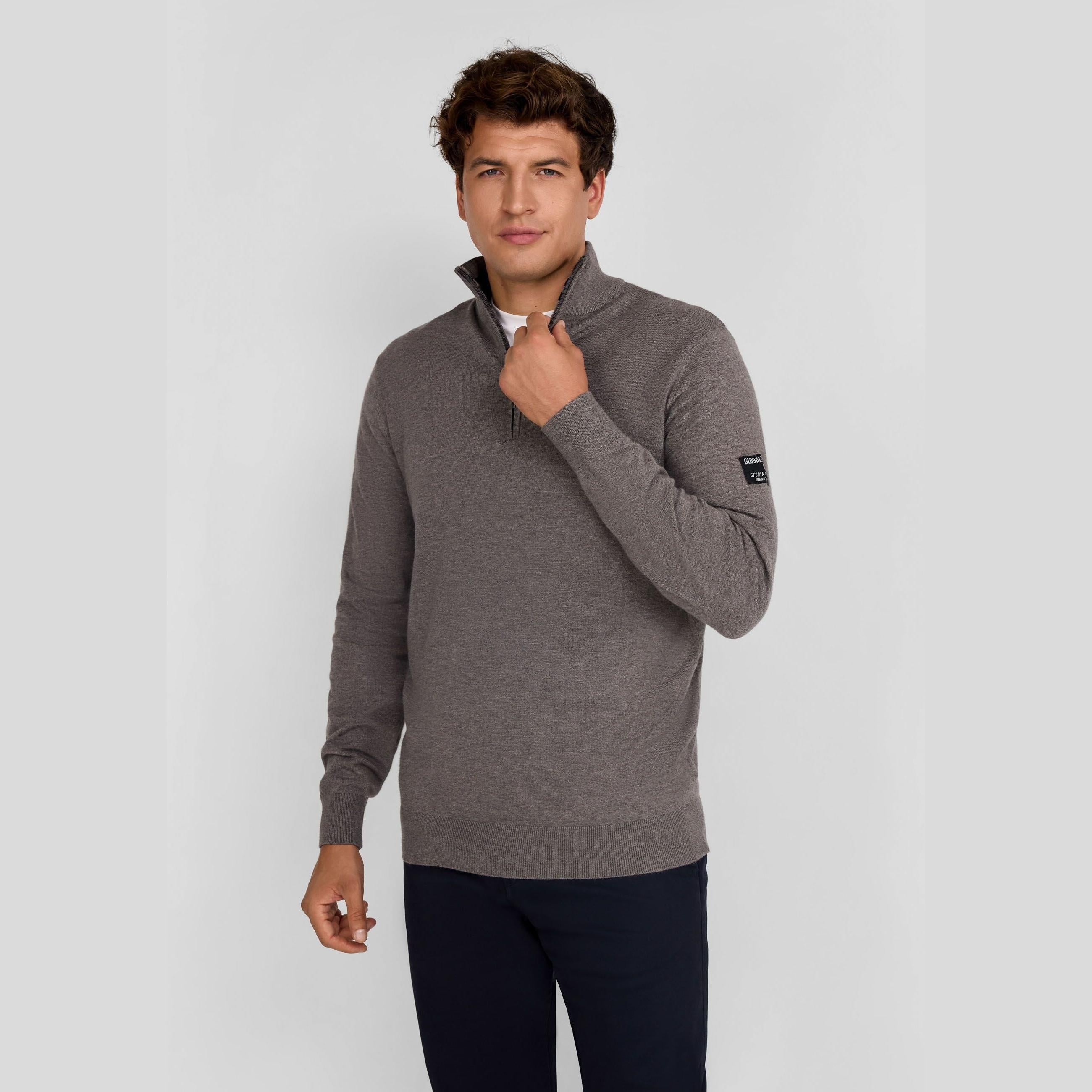 Young man wearing grey half zip jumper with stand collar, hand adjusting zip, paired with dark trousers against light background