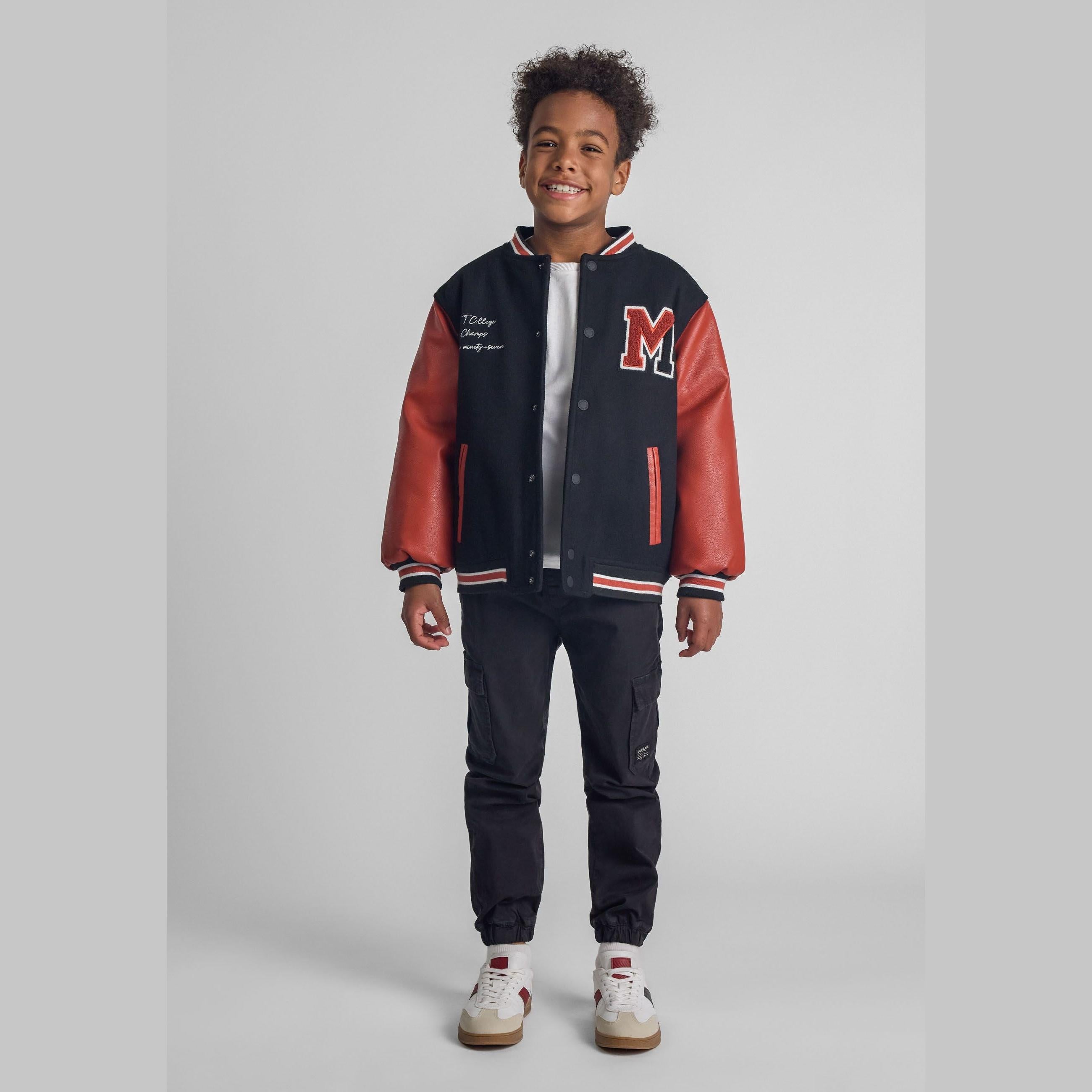 Young boy wearing black cargo trousers with red and navy varsity jacket, smiling against light grey studio background