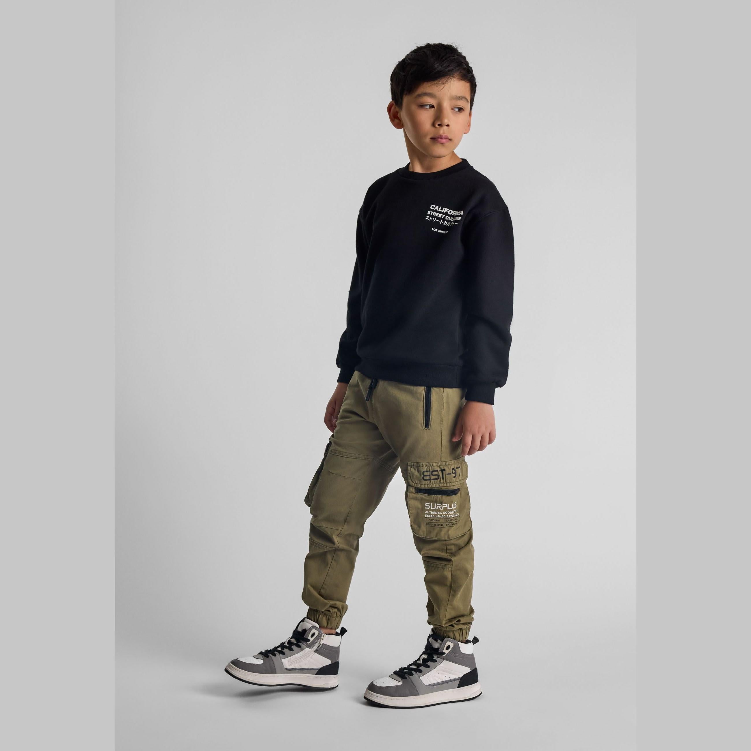 Young Asian boy wearing olive green cargo trousers with black sweatshirt and grey sneakers, standing against white studio background