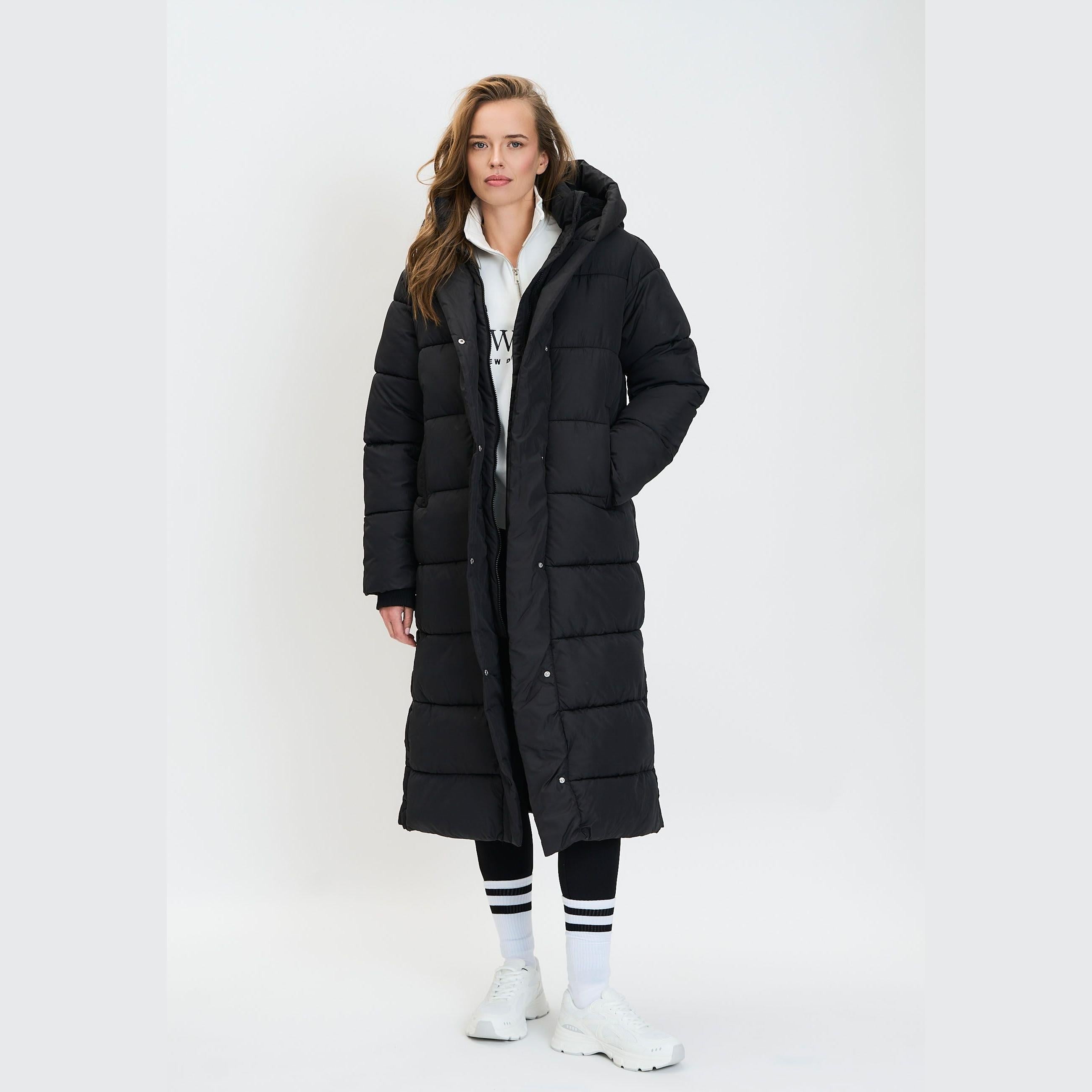 Model wearing long black quilted parka coat with hood up, paired with white hoodie underneath and striped socks, standing against white studio background