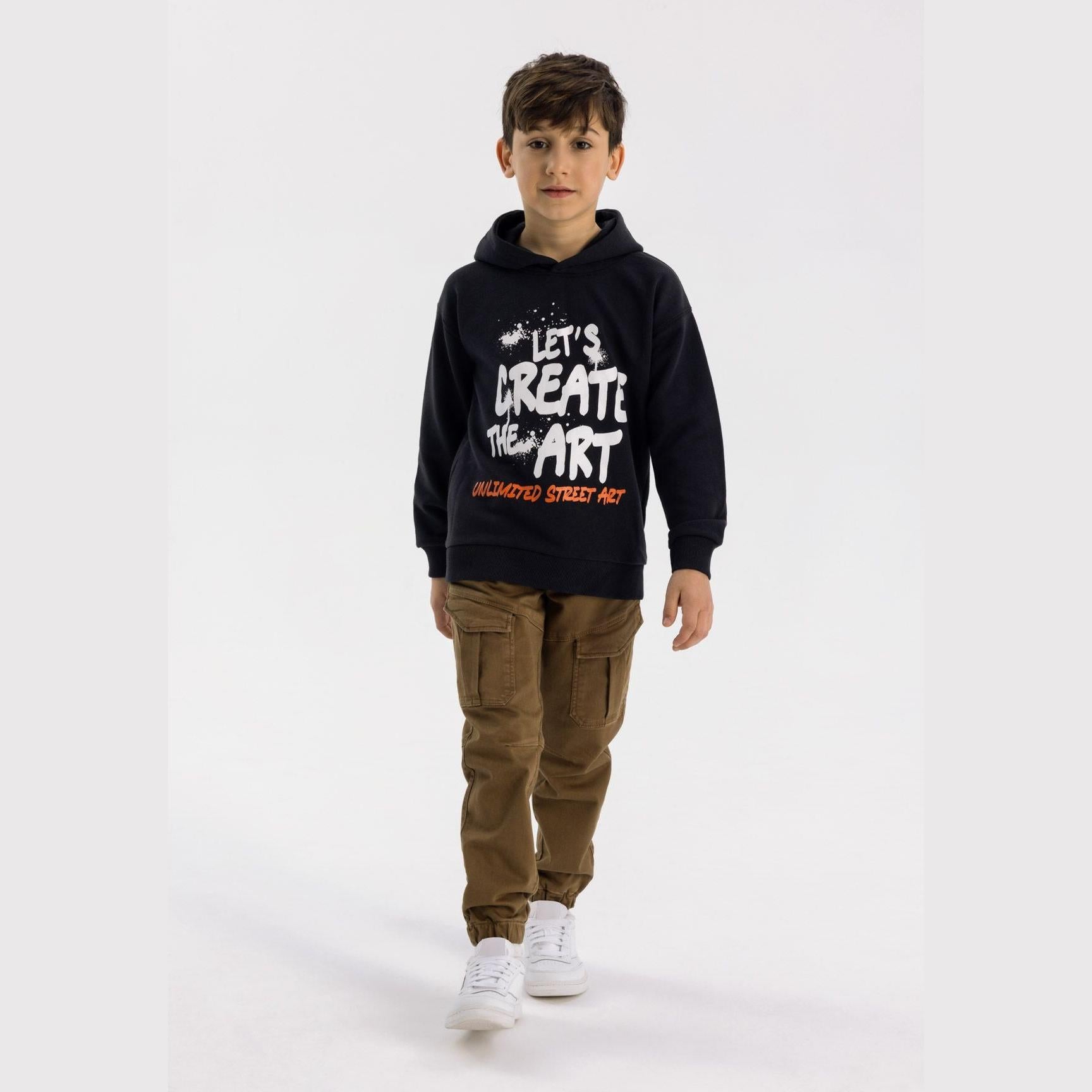 Boy wearing brown knitted cargo trousers with black hoodie against white background, showing full outfit styling