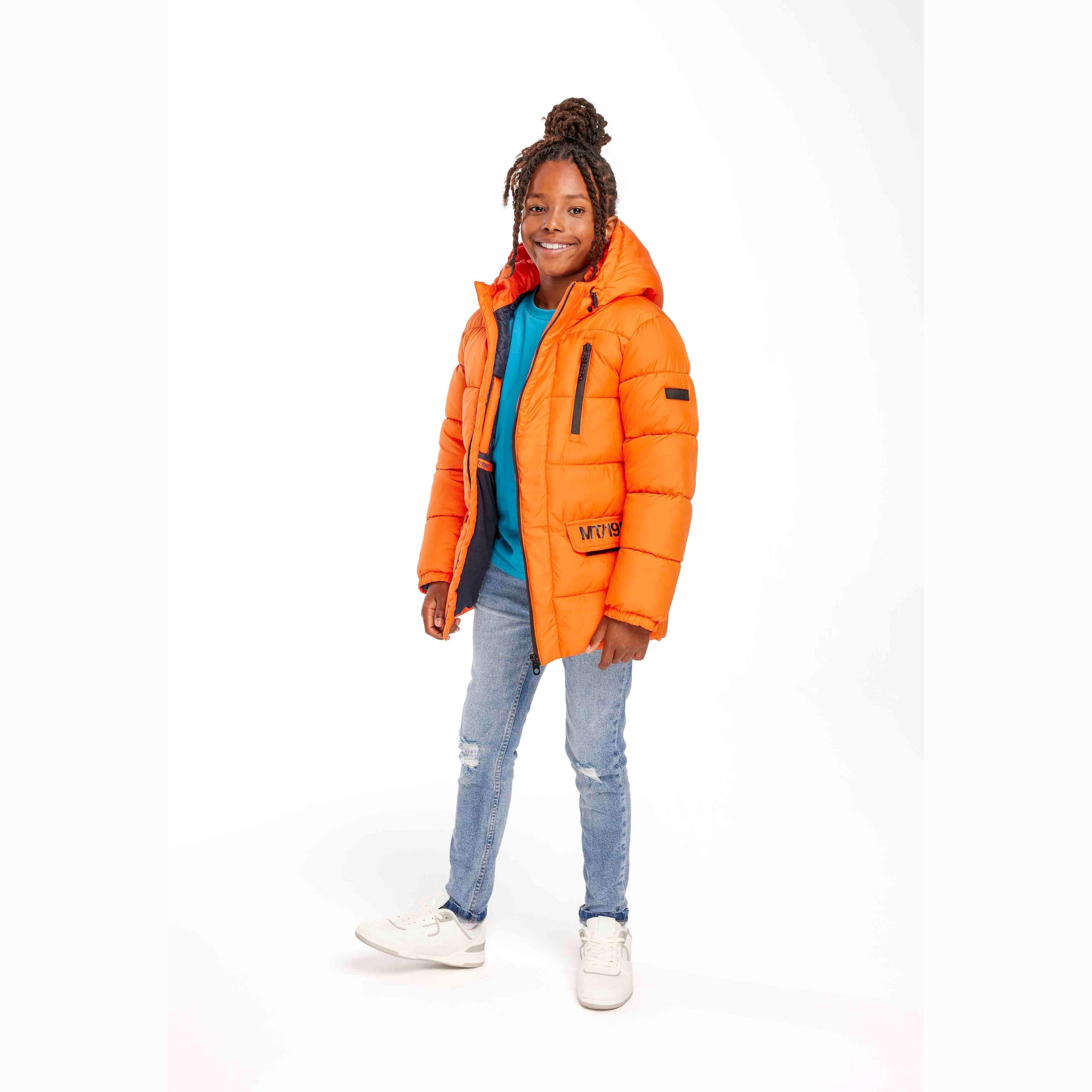 Young girl wearing bright orange padded winter jacket with hood and blue top, paired with distressed jeans and white sneakers, standing against white studio background