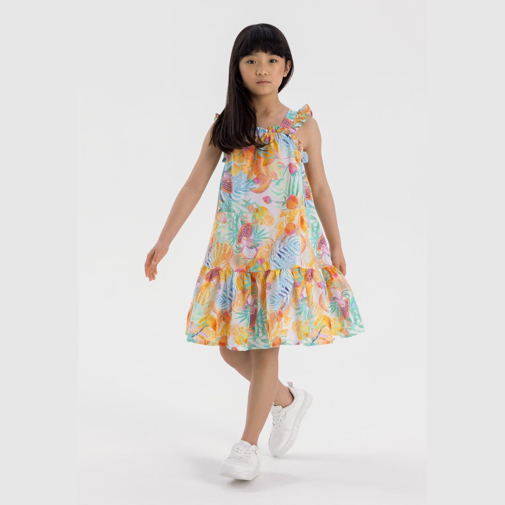 Young girl wearing tropical print cotton dress with tiered ruffles and shoulder straps, standing against white studio background wearing white sneakers
