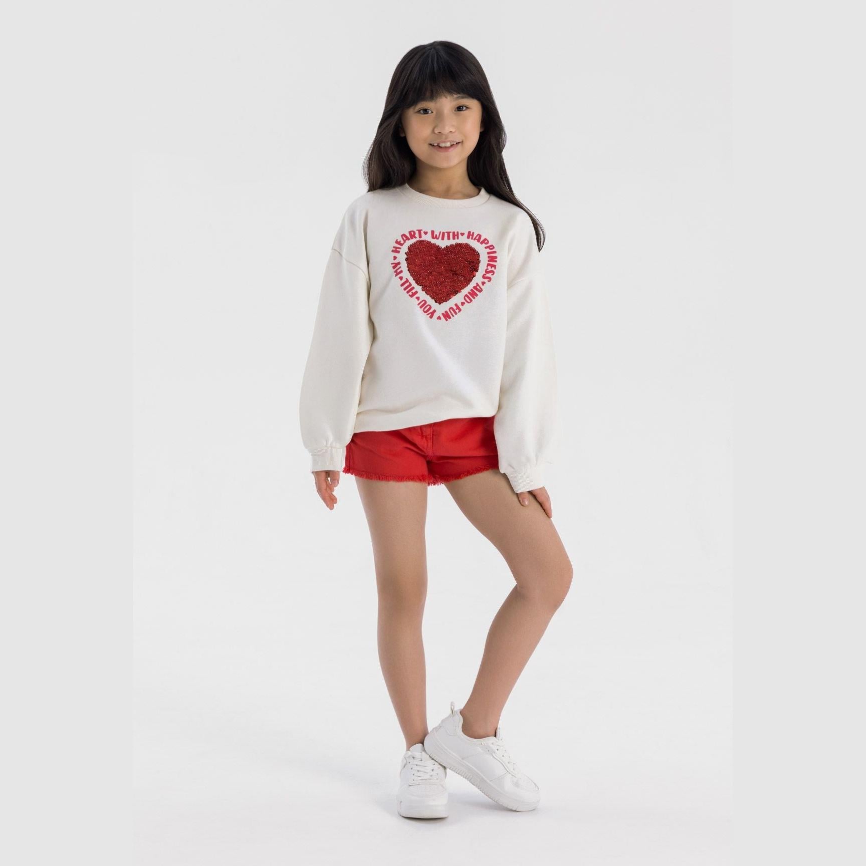 Young girl wearing cream fleece sweatshirt with red sequin heart design and red shorts, standing against white background