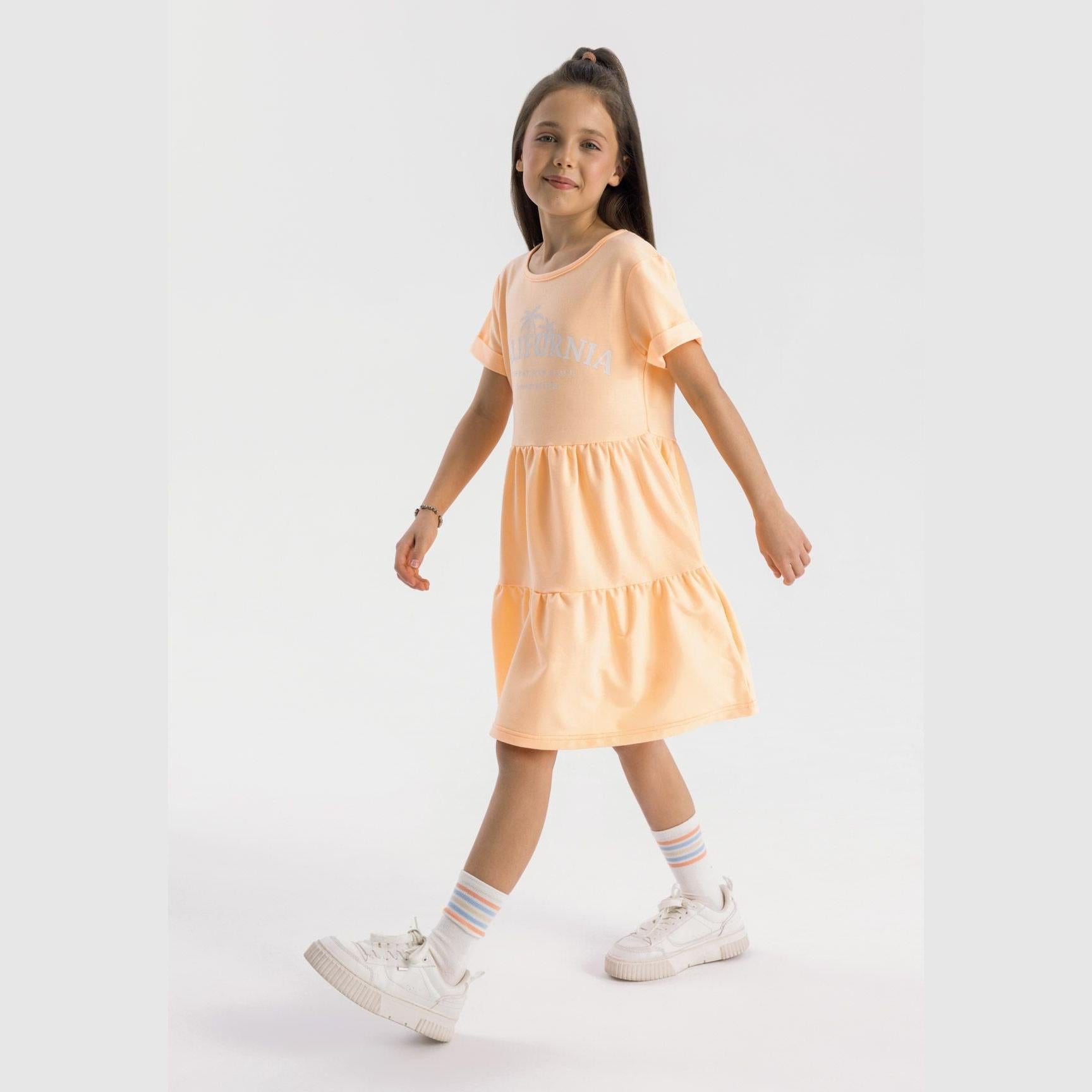 Girl wearing orange cotton fleece tiered dress with California palm print, styled with white sneakers and striped socks on white background