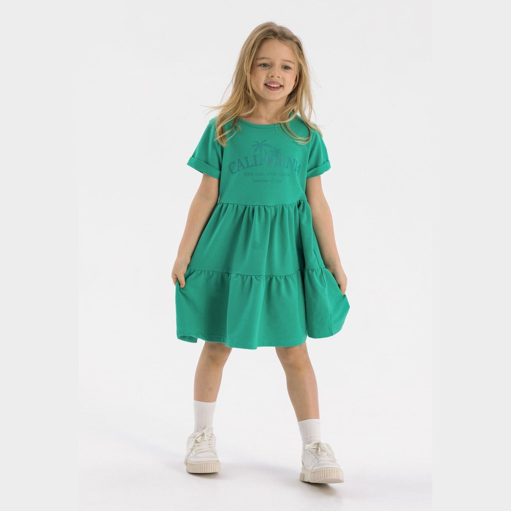 Young blonde girl wearing vibrant green tiered cotton dress with California print, standing against white studio background with white sneakers and socks