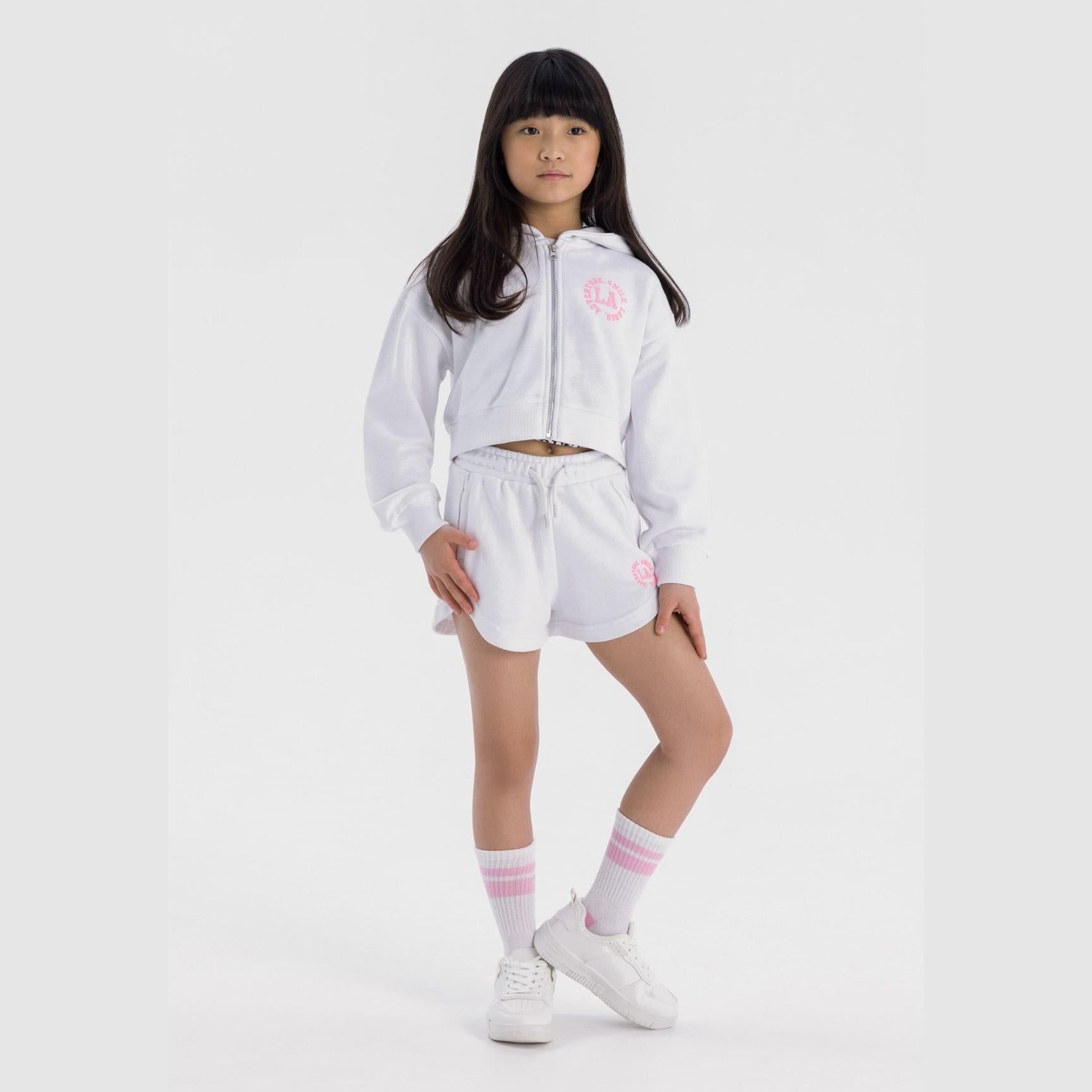 Girl wearing white zip hoodie and shorts set with pink L.A. branding, styled with white and pink striped socks and white sneakers on light grey background