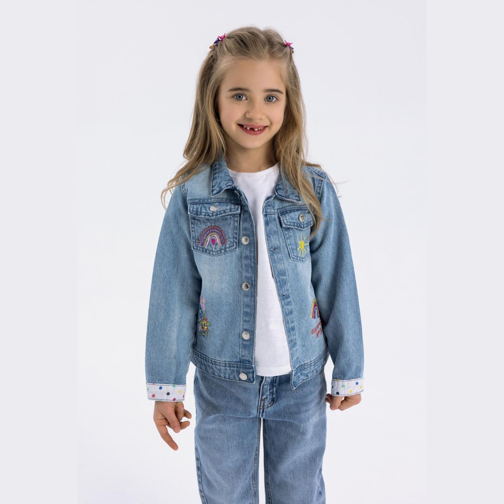Young girl wearing light blue embroidered denim jacket over white top with matching jeans, standing against white background
