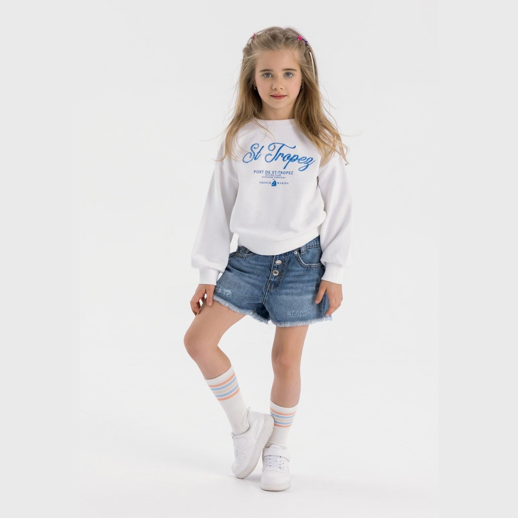 Young blonde girl wearing white St Tropez sweatshirt and navy denim shorts with frayed hems, styled with white sneakers and striped socks on white background