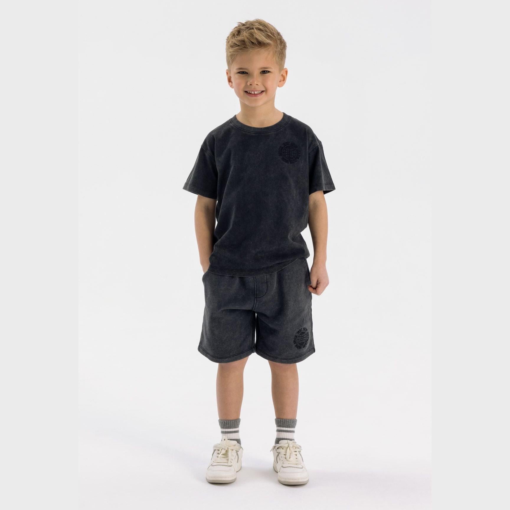 Young blonde boy wearing washed grey fleece t-shirt and matching shorts set, standing against white background with hands in pockets, wearing white sneakers and striped socks