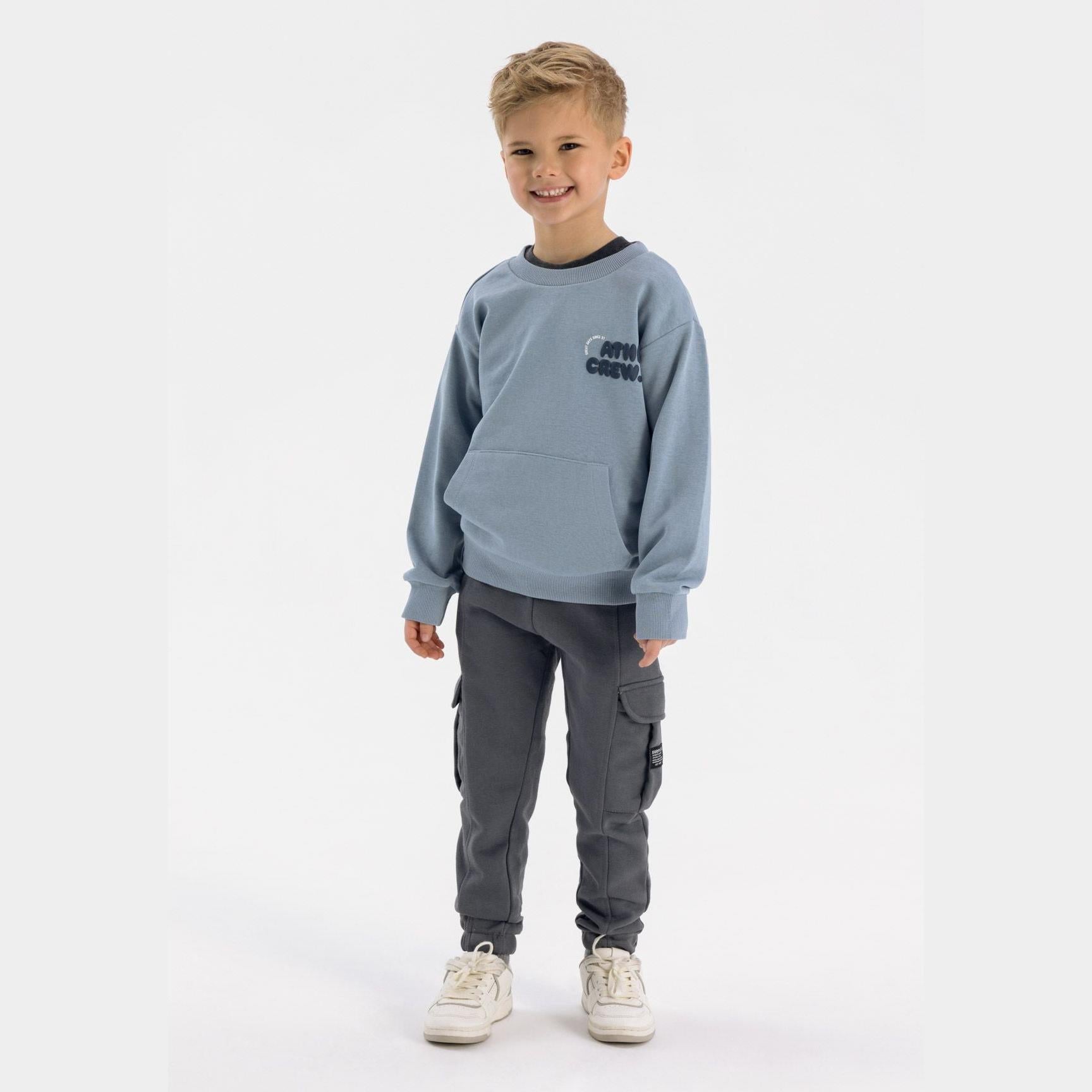 Young boy wearing grey cargo joggers with blue sweatshirt, standing against white background, full body view showing complete outfit styling