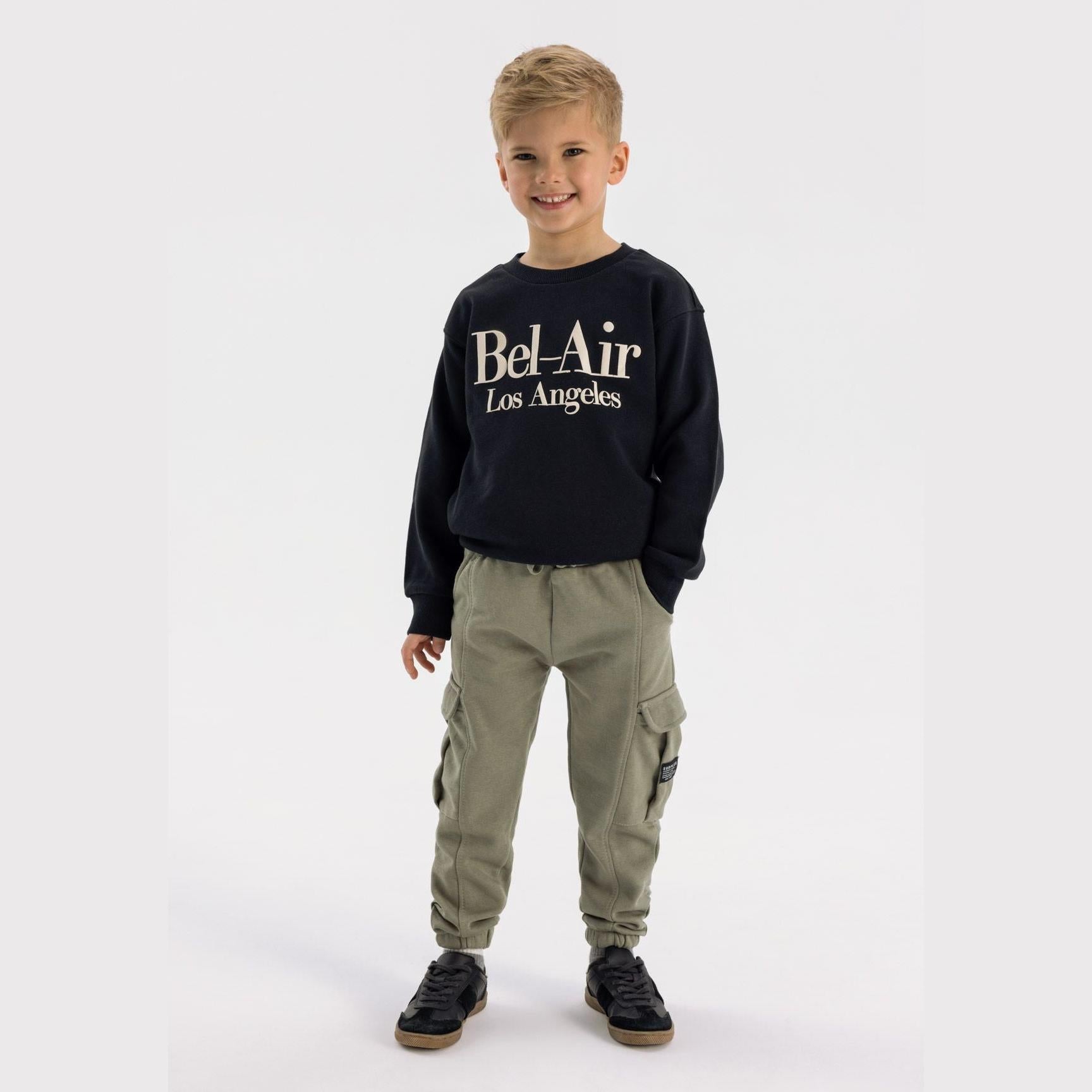 Young boy wearing black Bel-Air Los Angeles sweatshirt and khaki cargo joggers with side pockets, standing against white background