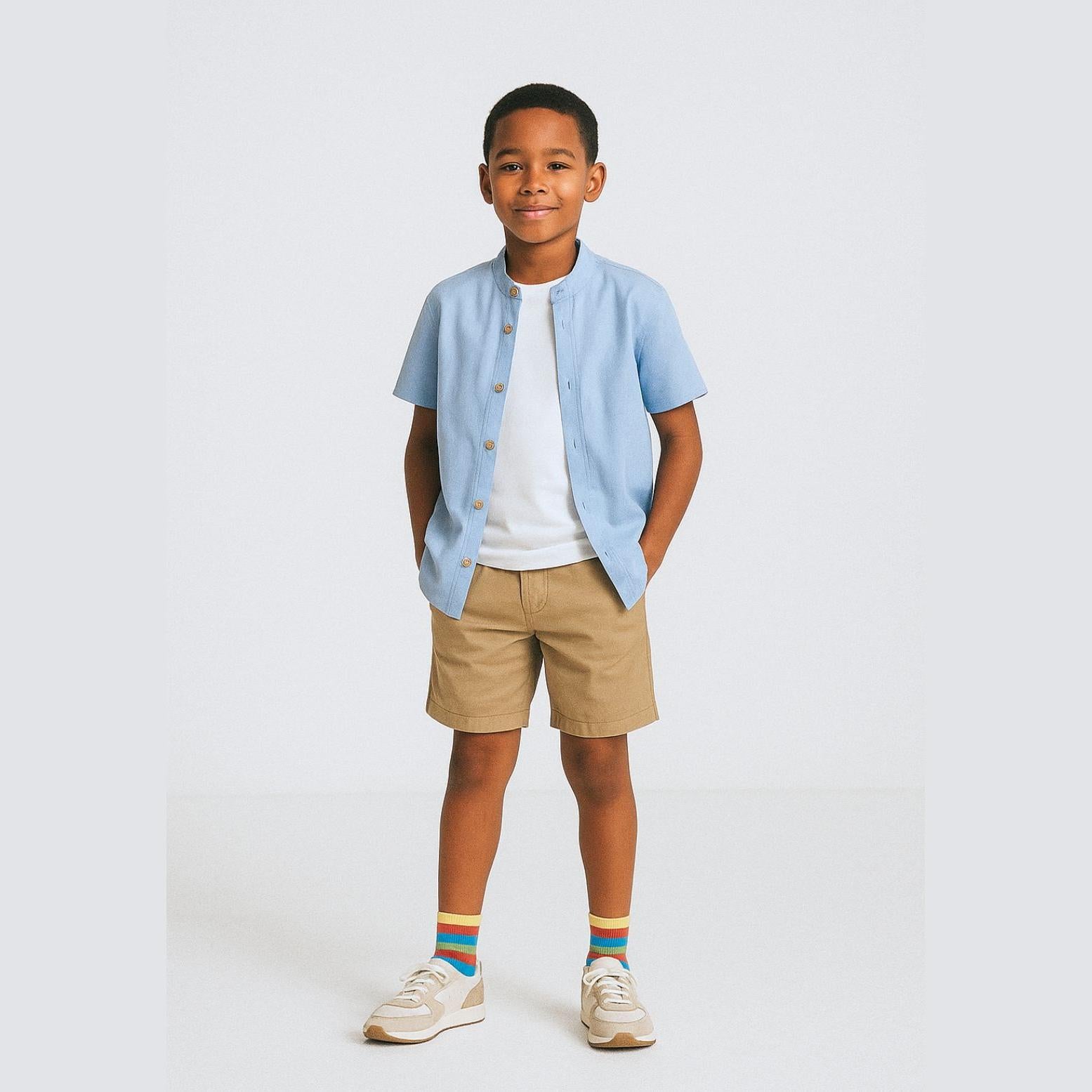 Boy wearing light blue grandad collar shirt and stone beige chino shorts set, standing against white background with hands in pockets