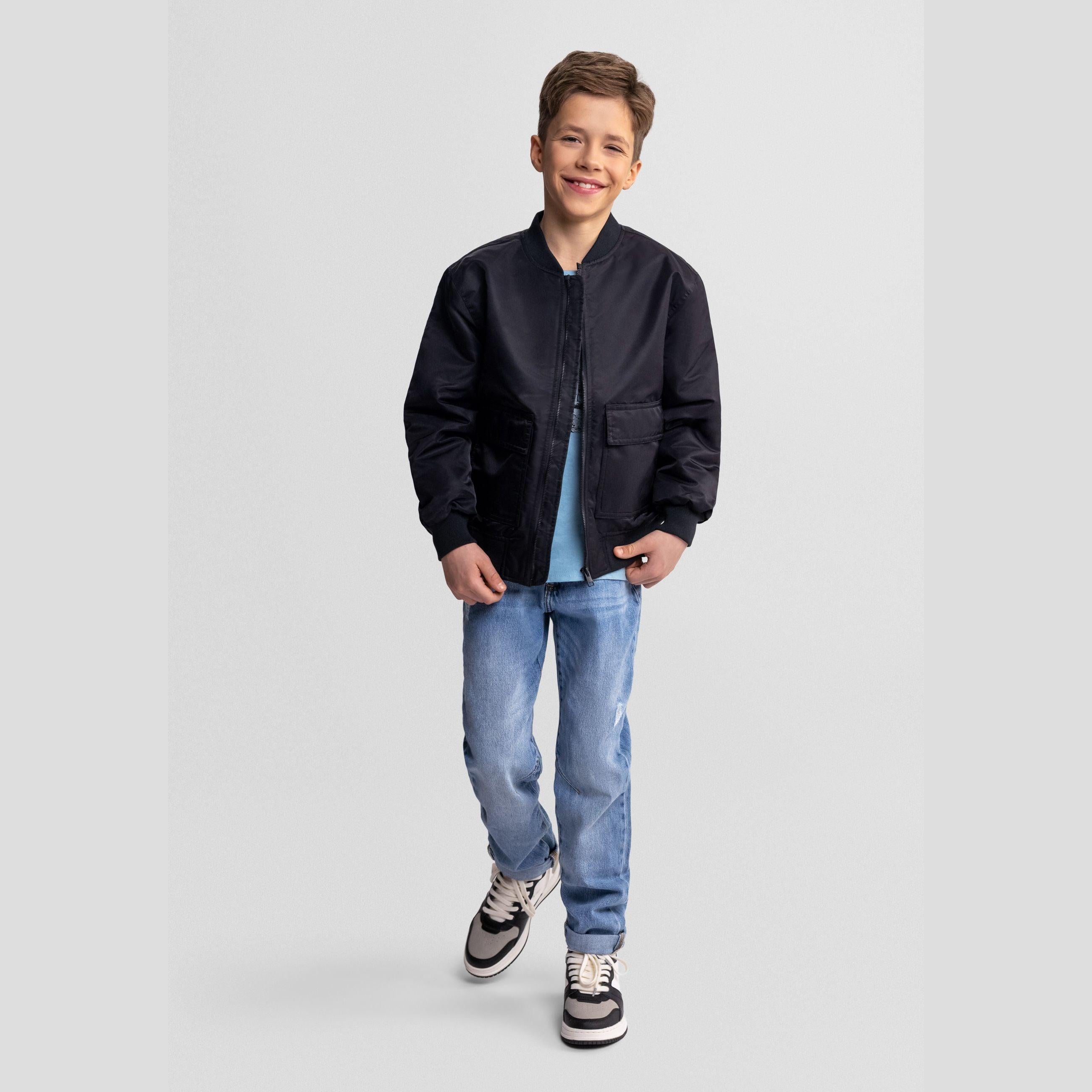 Young boy wearing black bomber jacket with light blue t-shirt and jeans, smiling against light background