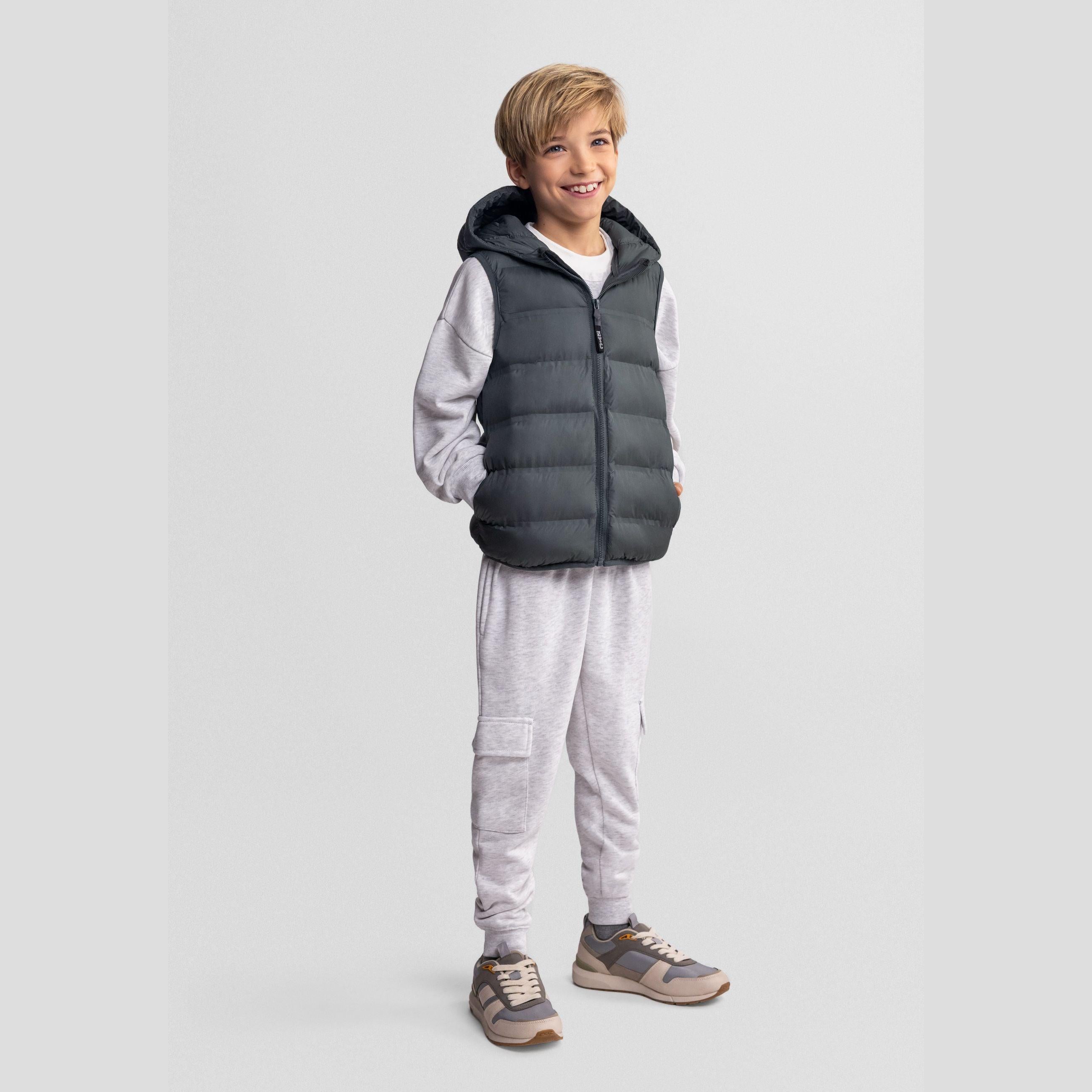 Young blonde boy wearing olive quilted hooded waistcoat over grey hoodie and joggers, smiling at camera against white background