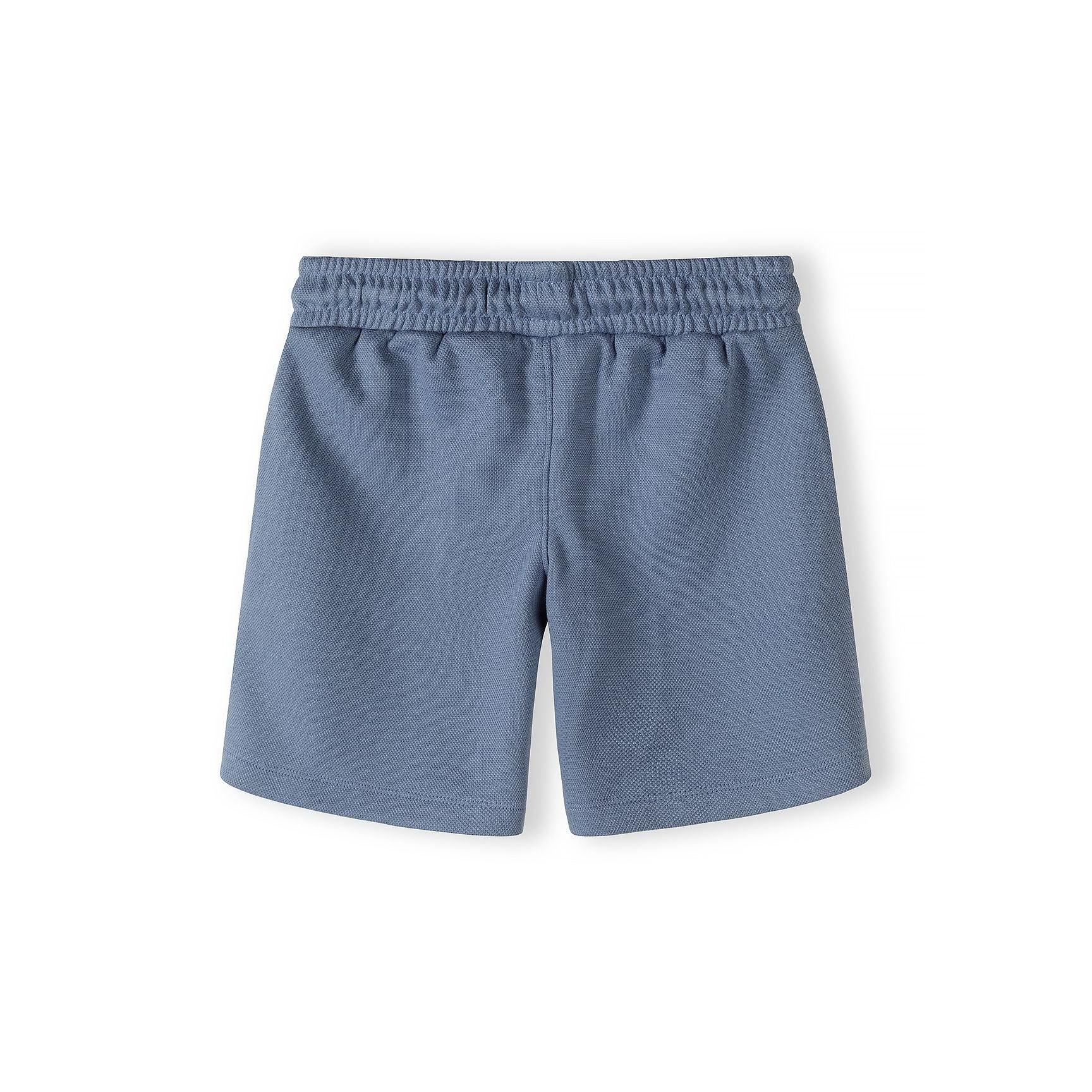 Close-up detail of blue polo shirt showing zip collar, embroidered Minoti logo, and pique texture with brand label visible