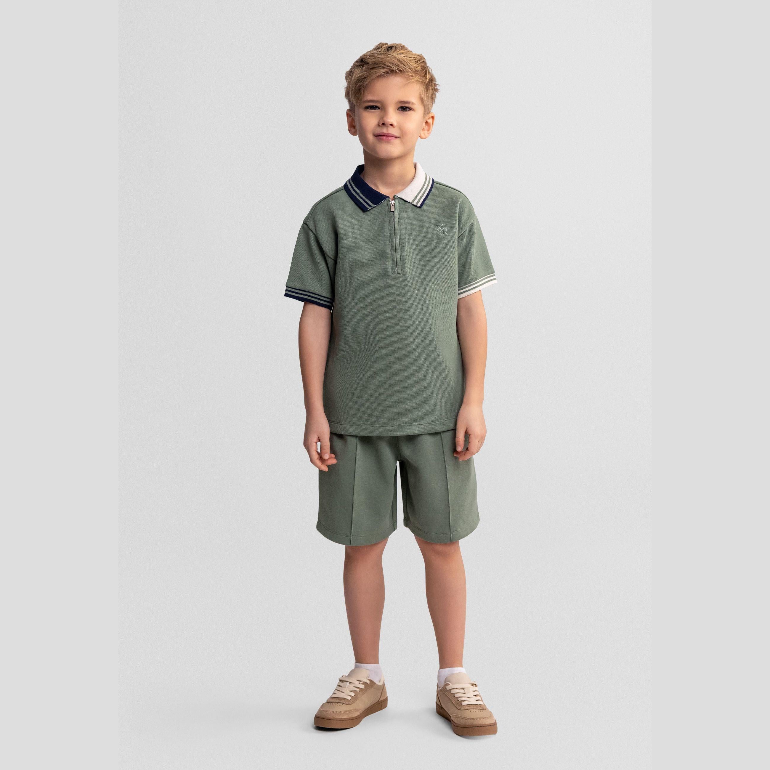 Young boy wearing teal pique polo shirt and matching shorts set, standing against light grey background, brown sneakers