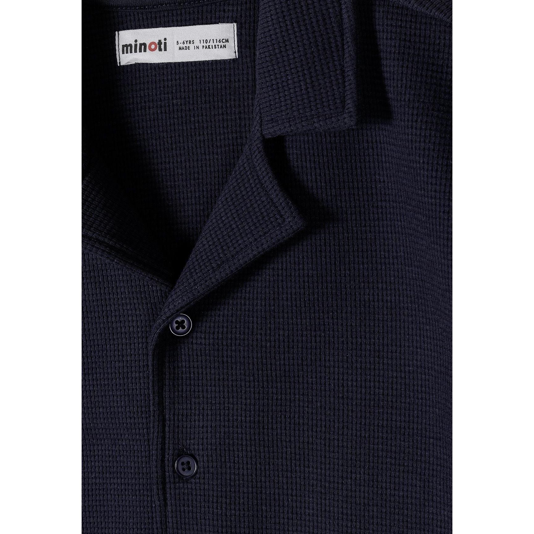 Close-up detail of navy polo shirt collar and button placket showing Minoti label and textured cotton fabric
