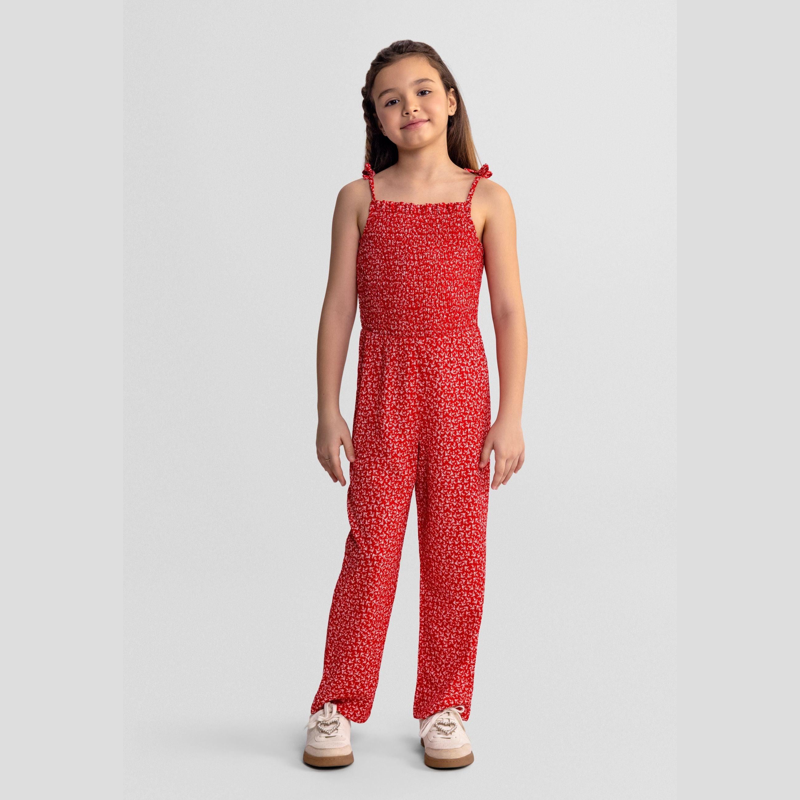 Girl wearing red and white floral jumpsuit with shirred bodice, tie straps, standing against grey background