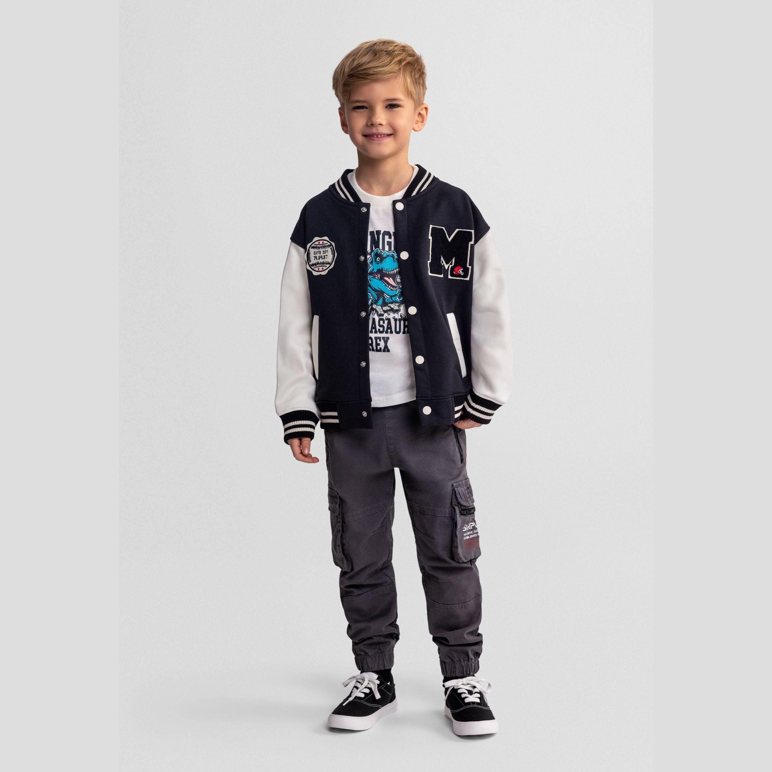 Young boy wearing black baseball jacket with white sleeves, white t-shirt and grey cargo pants, standing against light grey background