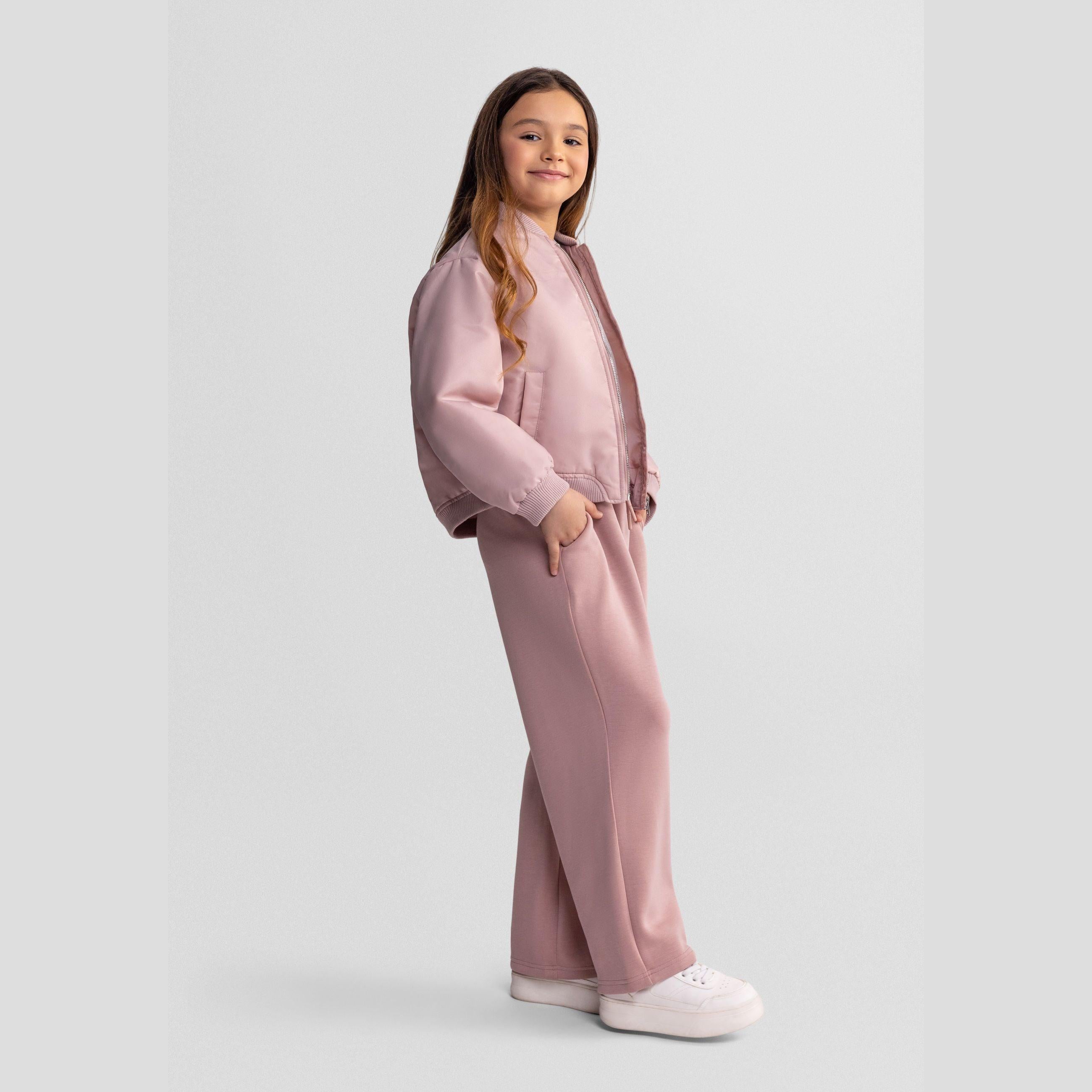 Side view of girl in pink quilted bomber jacket and matching trousers, white sneakers, showing profile against white studio background