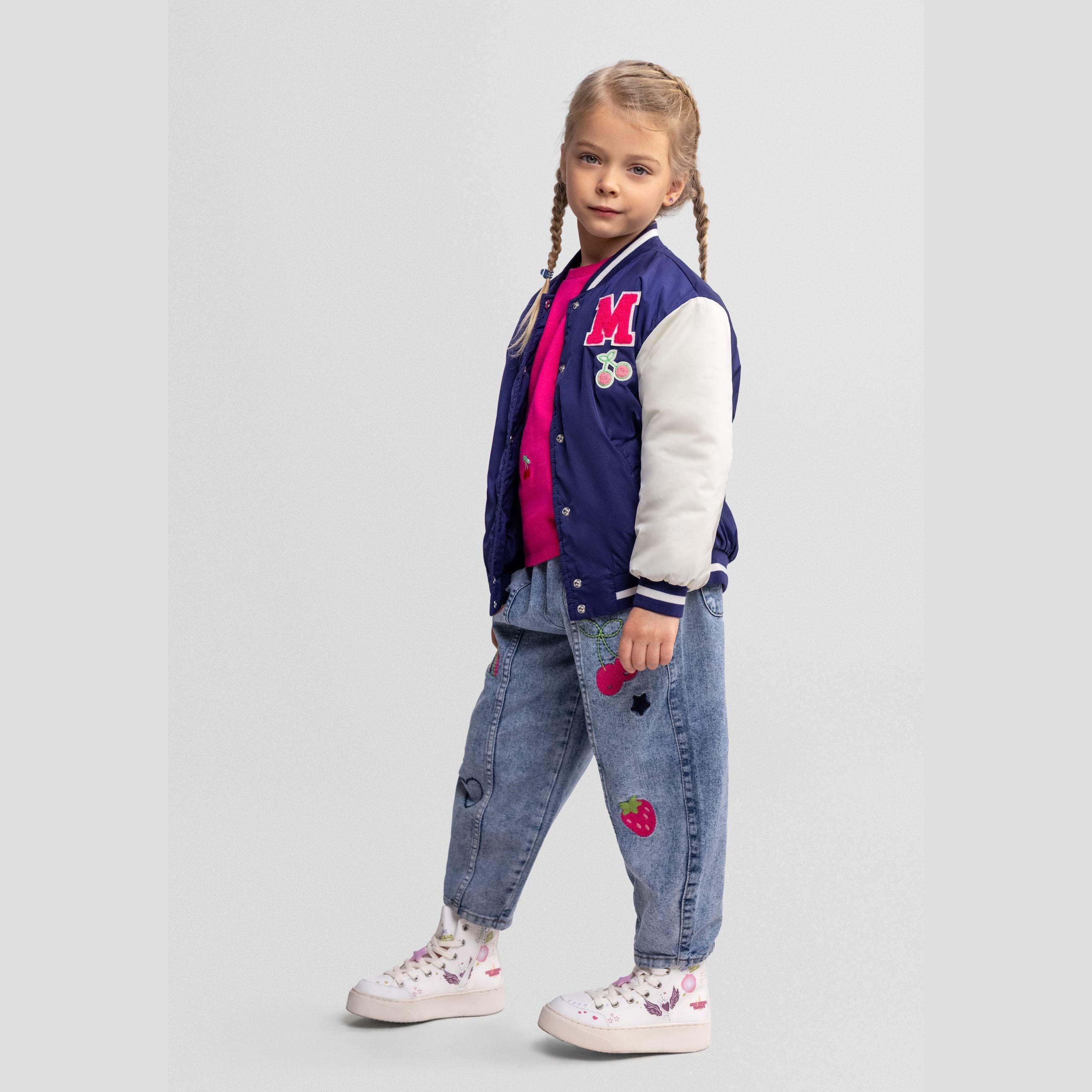 Young girl wearing navy and white varsity bomber jacket with pink top and embroidered jeans, standing against light grey background