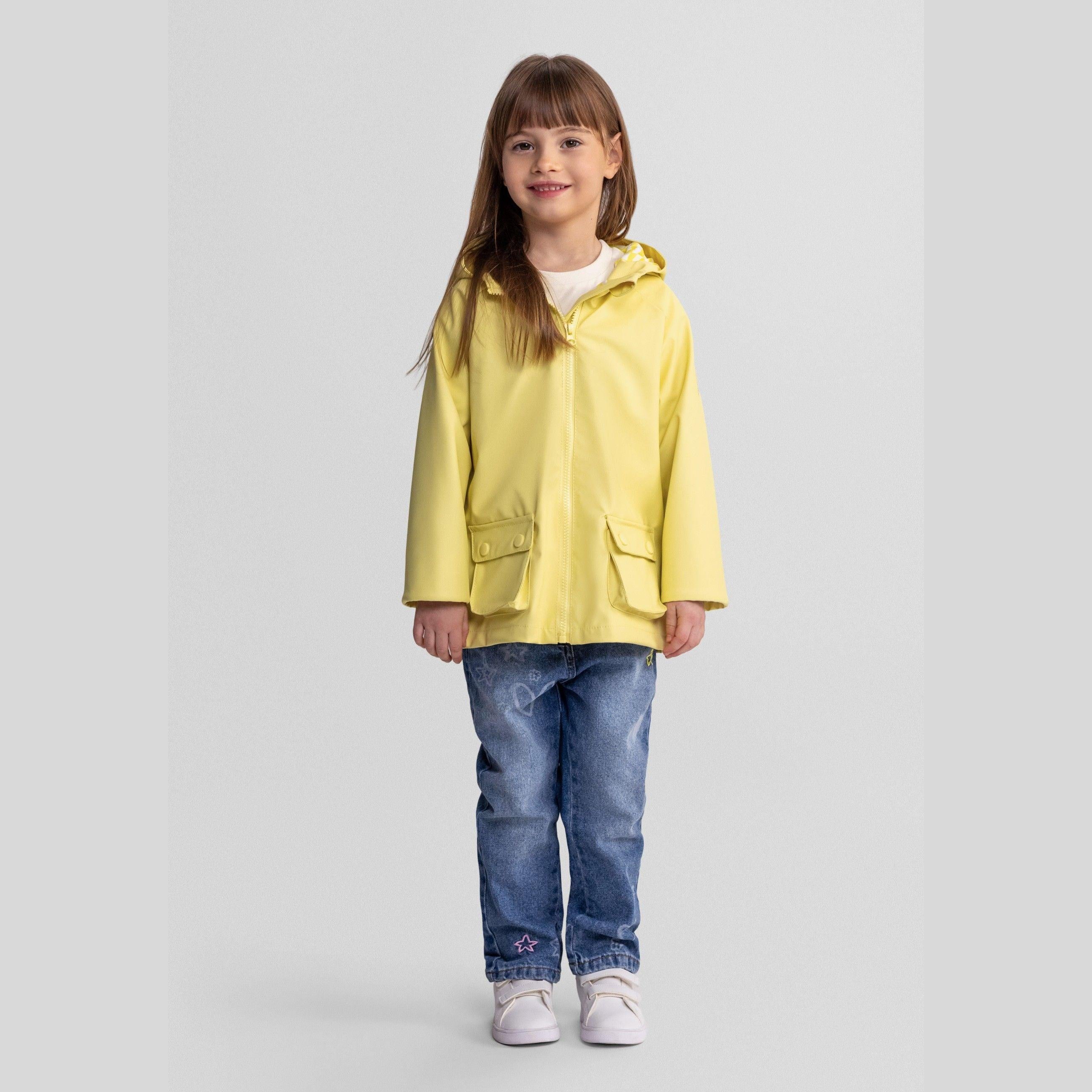 Young girl wearing bright yellow waterproof rain jacket with hood and blue jeans, standing against white background, smiling