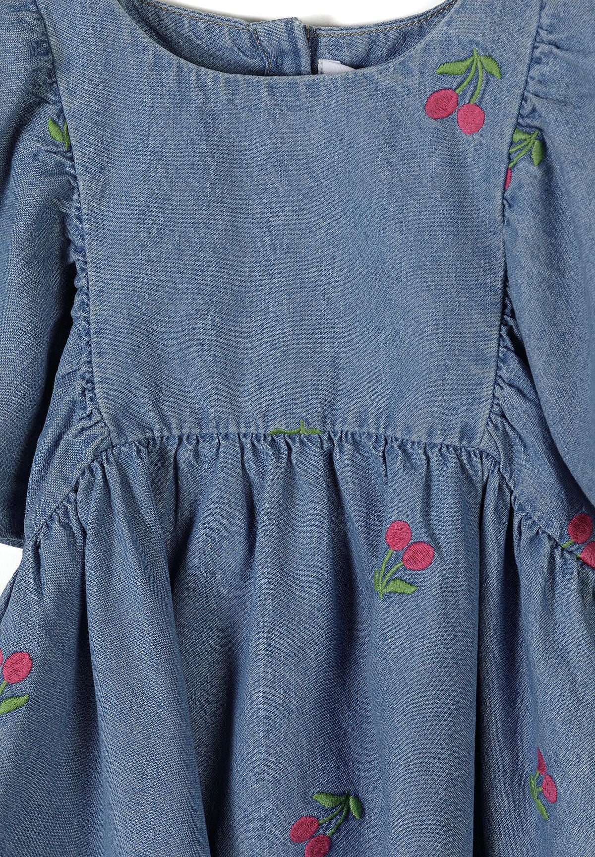 Close-up detail of blue denim dress showing cherry embroidery, gathered waist seam and fabric texture