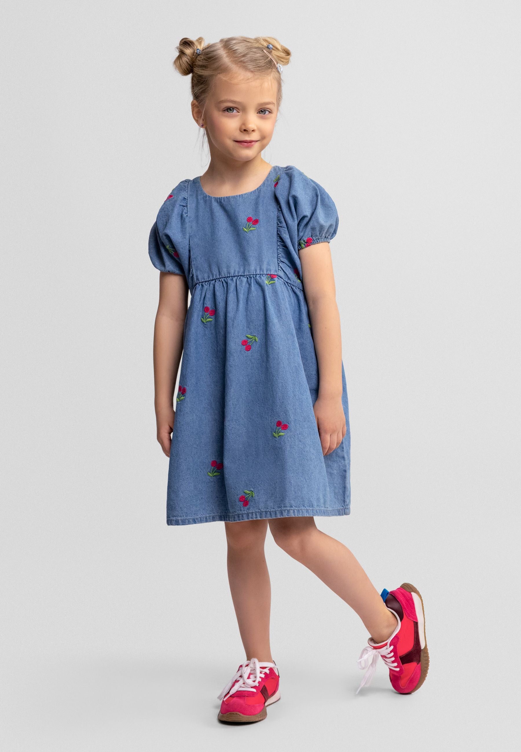 Young girl wearing blue denim dress with cherry embroidery, posed against light grey background, wearing pink trainers