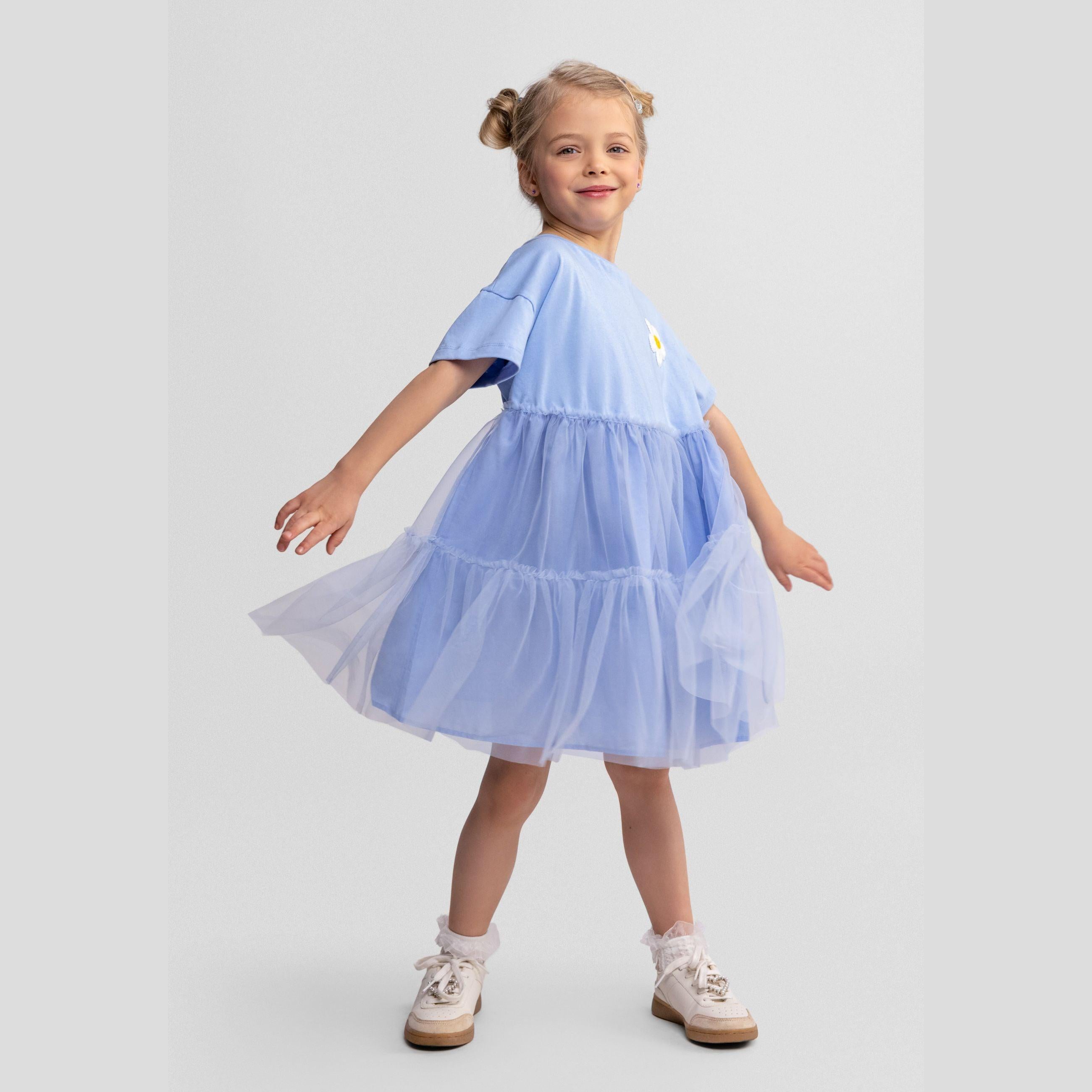 Young girl wearing blue cotton and tulle dress with embroidered daisy, twirling against white studio background