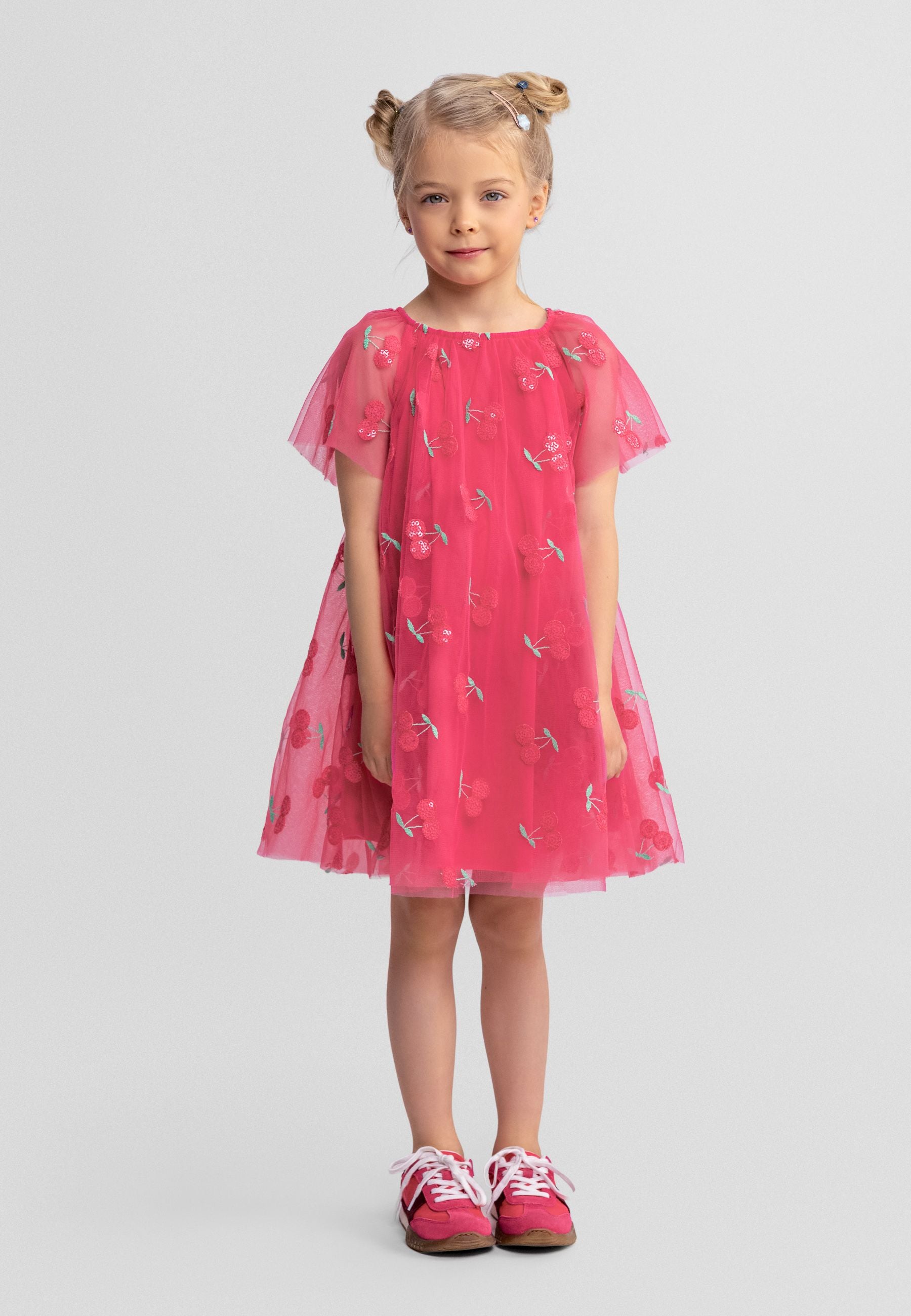 Young blonde girl wearing pink tulle dress with sequin cherry pattern and angel sleeves, standing against white background wearing pink shoes