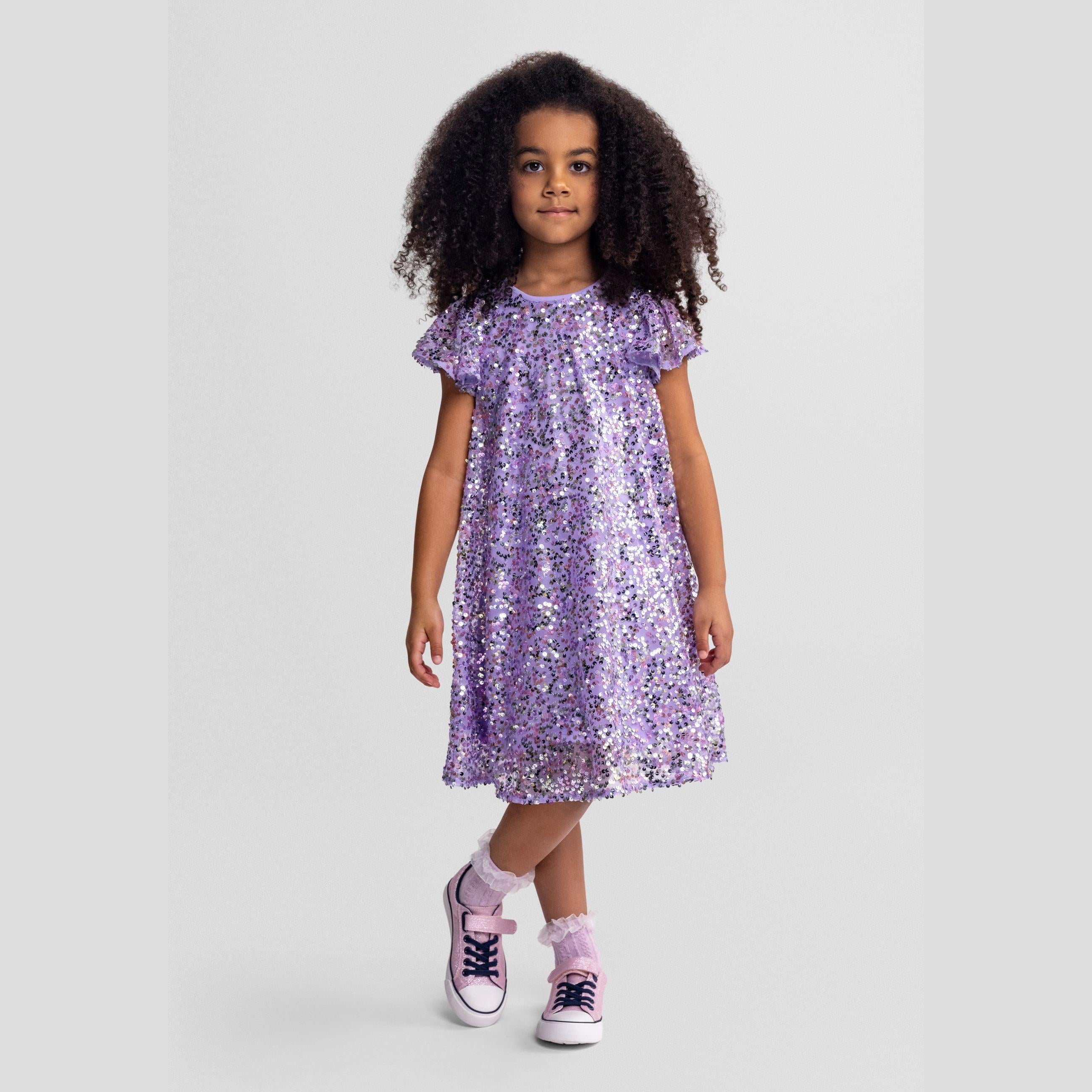 Girl wearing purple sequin party dress with multicolour sparkles, A-line silhouette and short flared sleeves, standing against light grey background