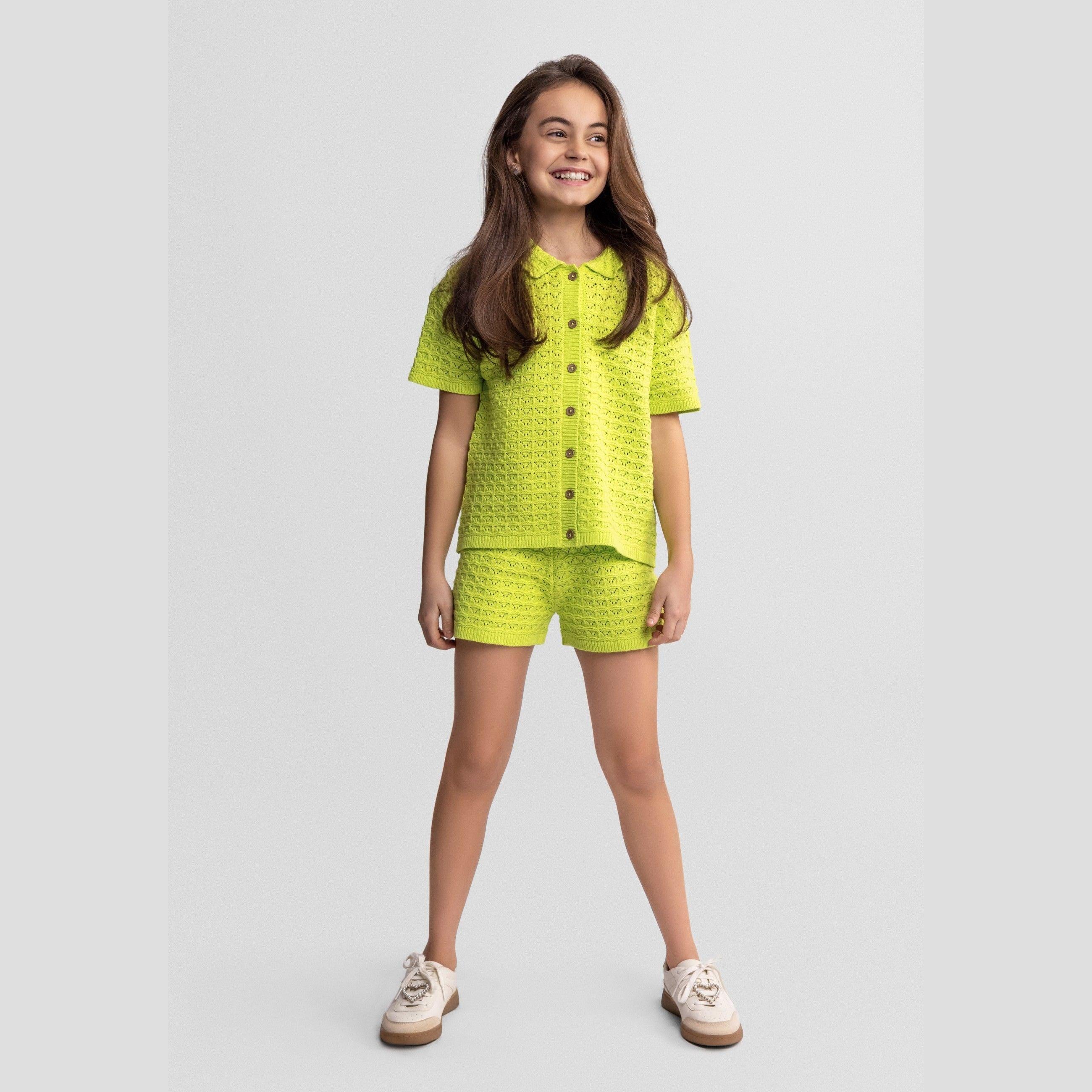 Young girl wearing lime green pointelle knit set with button-front top and matching shorts, standing against white background in casual pose