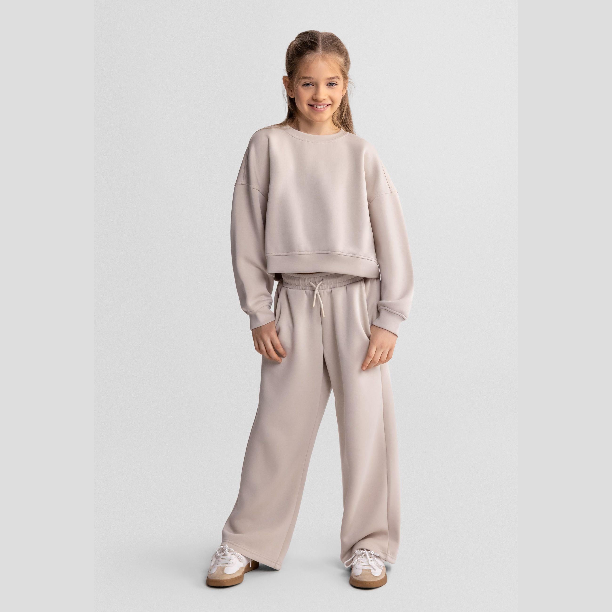 Girl wearing beige wide leg joggers with matching sweatshirt, standing against white background, showing full outfit coordination