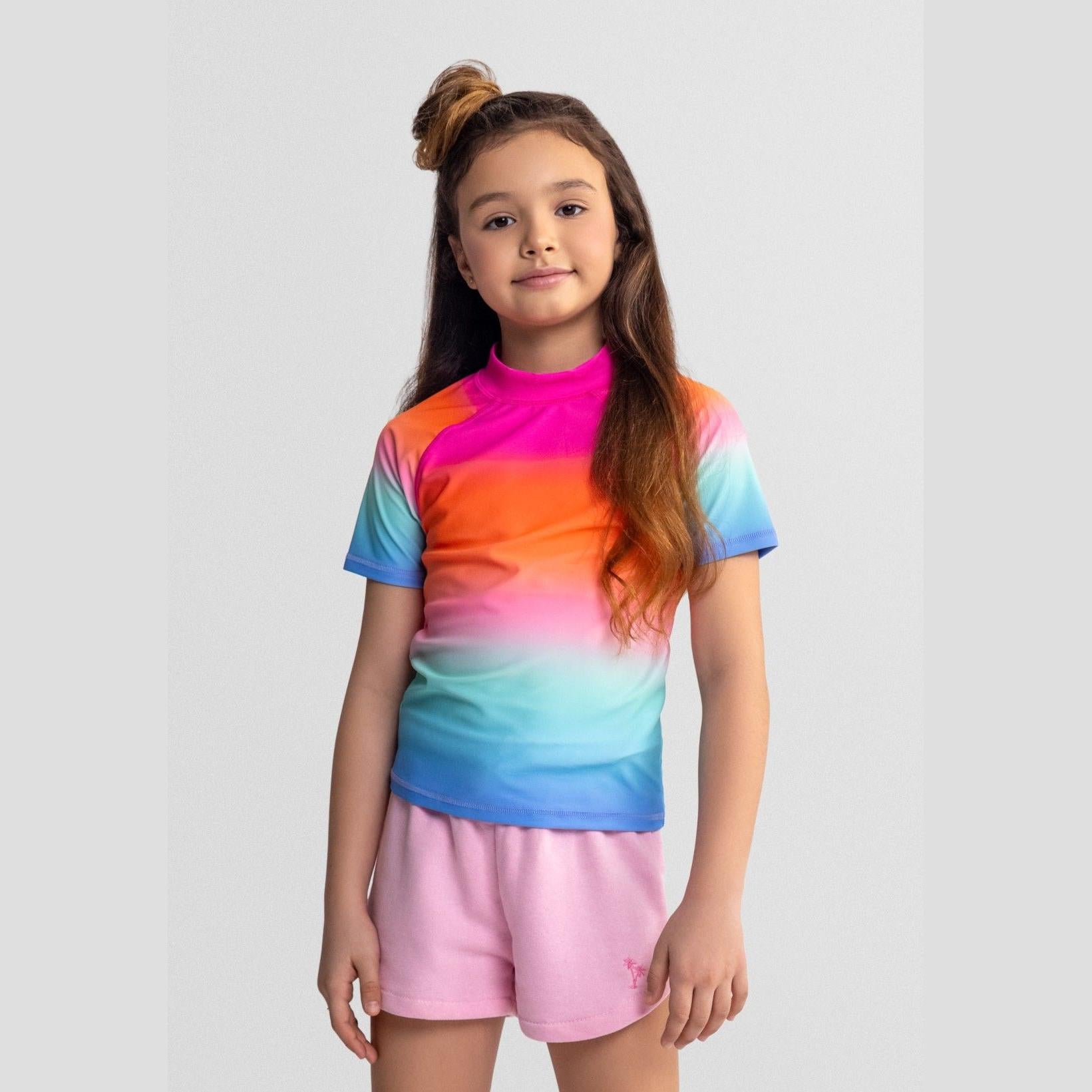 Young girl wearing rainbow ombre rash top with pink shorts, smiling against neutral background