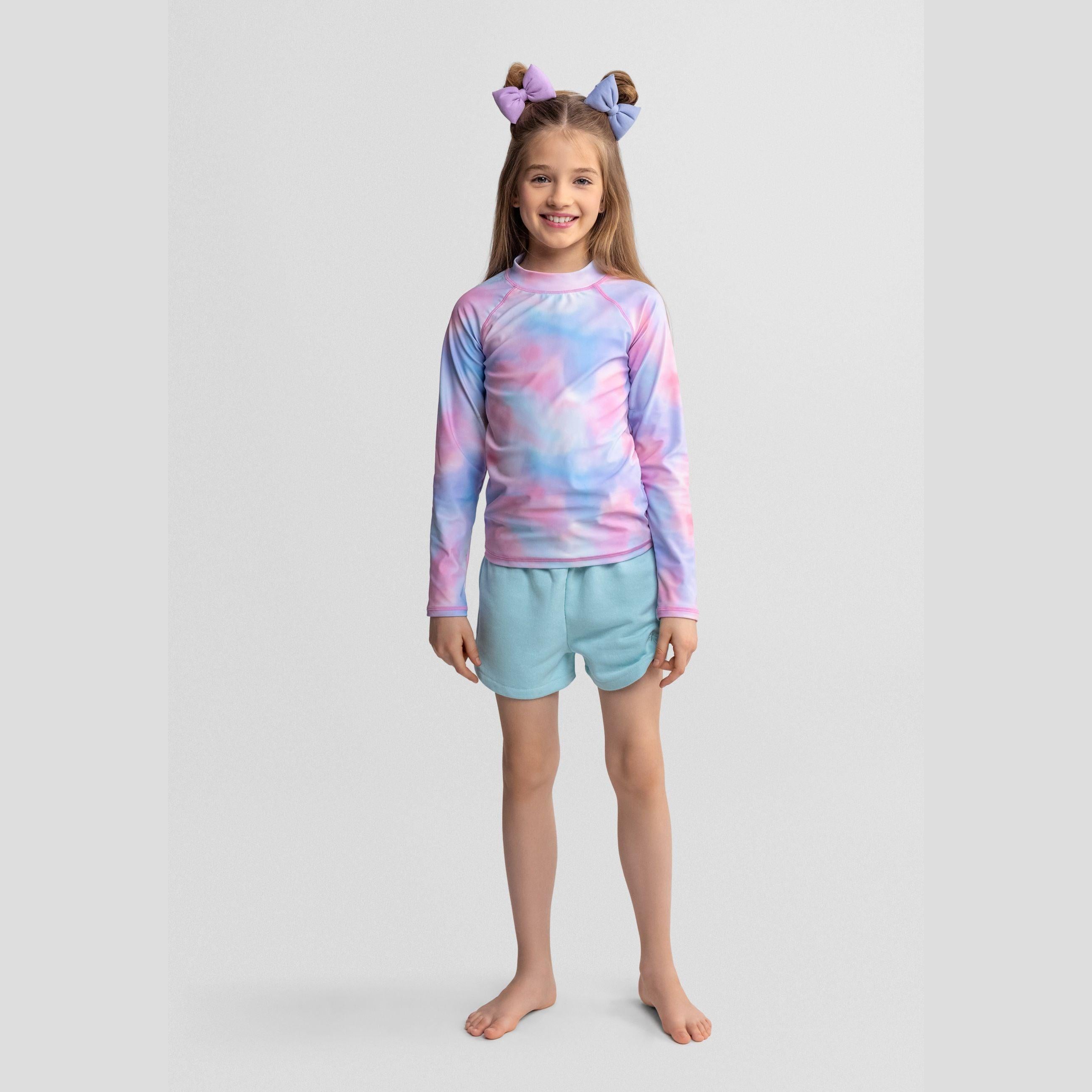 Young girl wearing pastel tie-dye long sleeve swim t-shirt with mint green shorts and matching hair bows, standing against white background