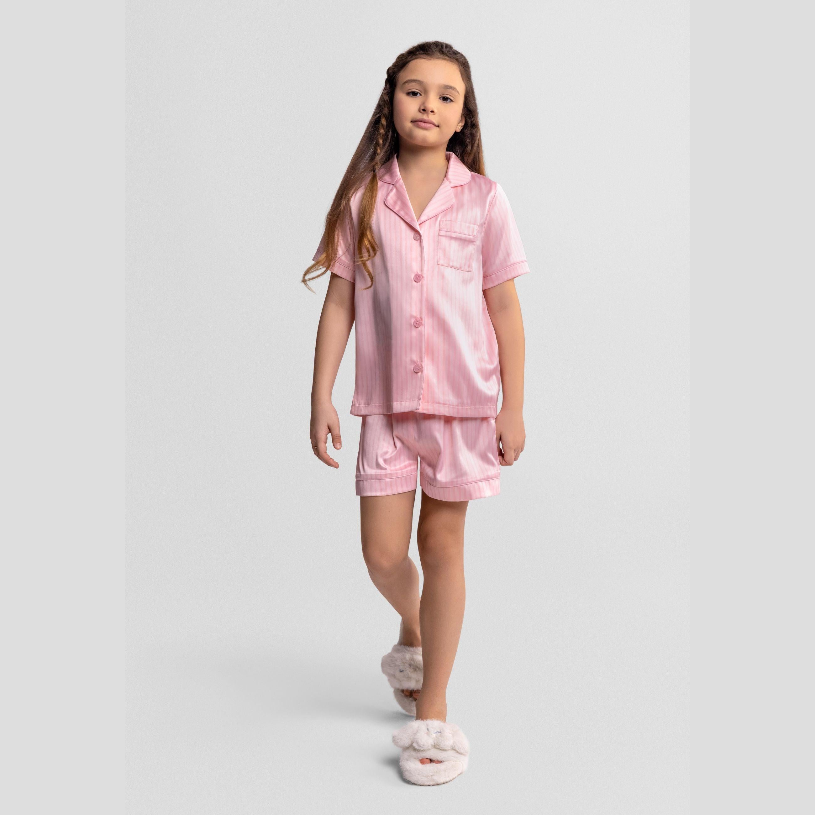 Girl wearing pink and white striped satin pyjama set with button-front top and shorts, standing against light grey background