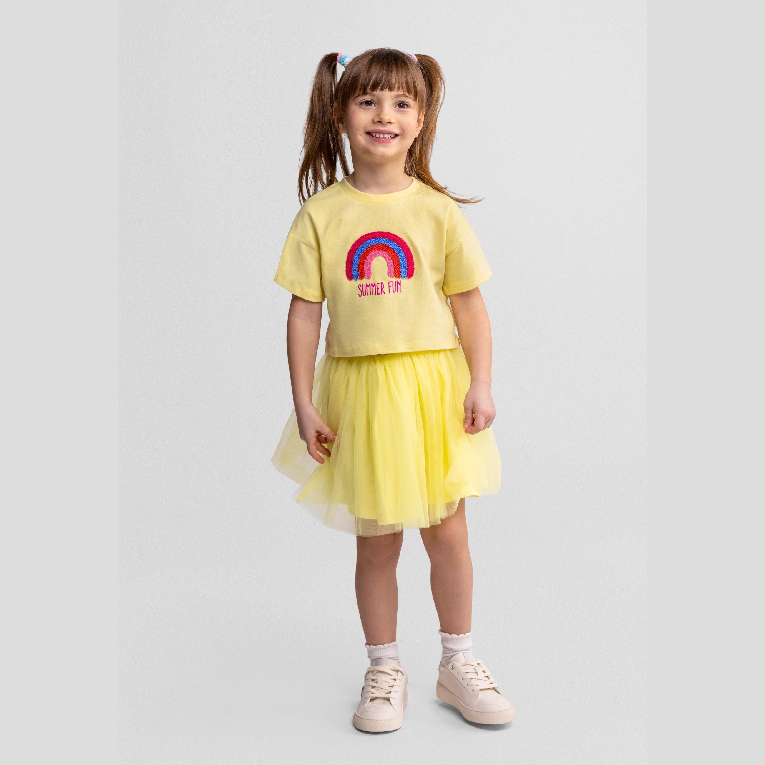 Young girl wearing pale yellow cotton t-shirt with rainbow applique and matching yellow tulle skirt, standing against white background