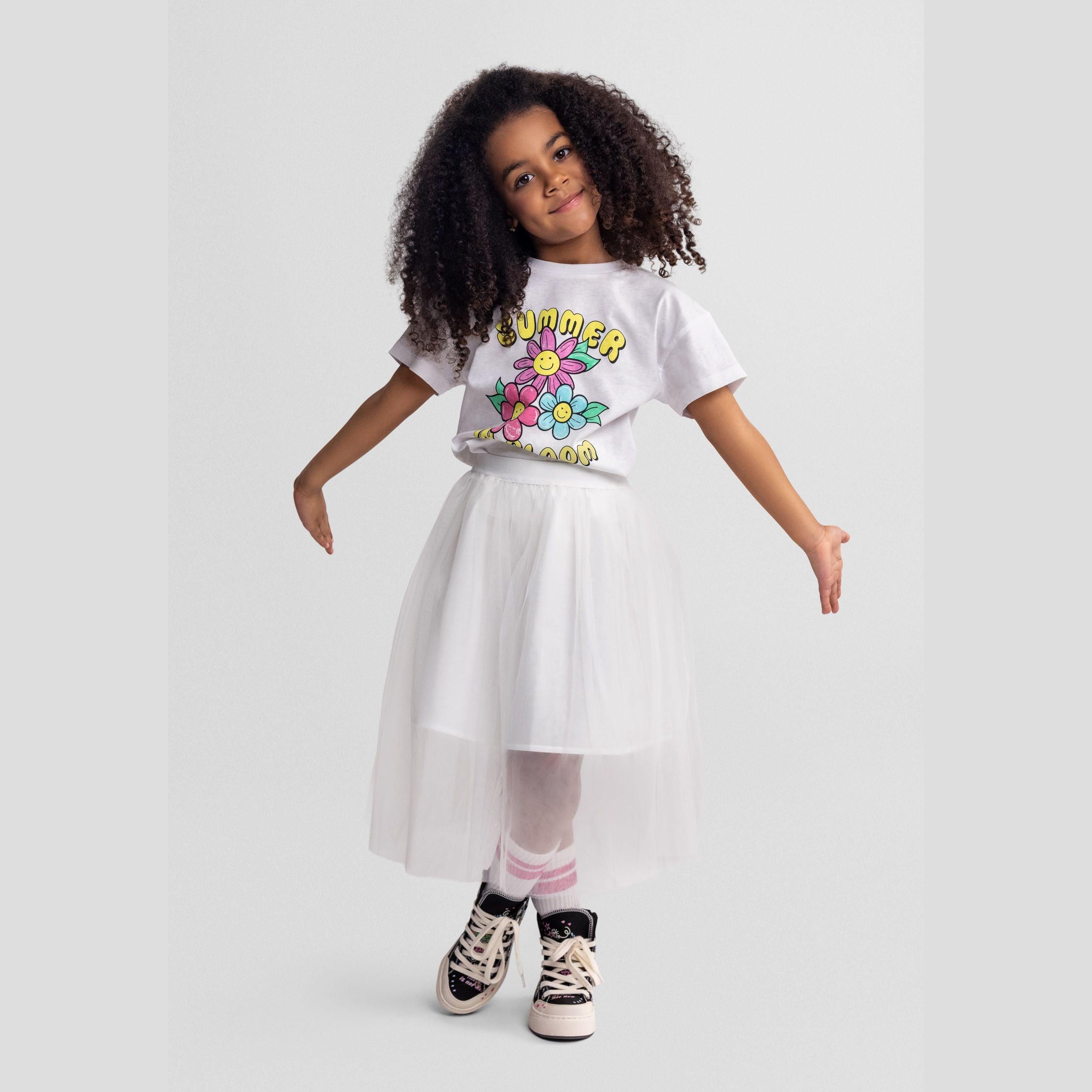 Young model wearing white t-shirt with Summer in Bloom floral print and white tulle skirt, styled with striped socks and sneakers against grey background
