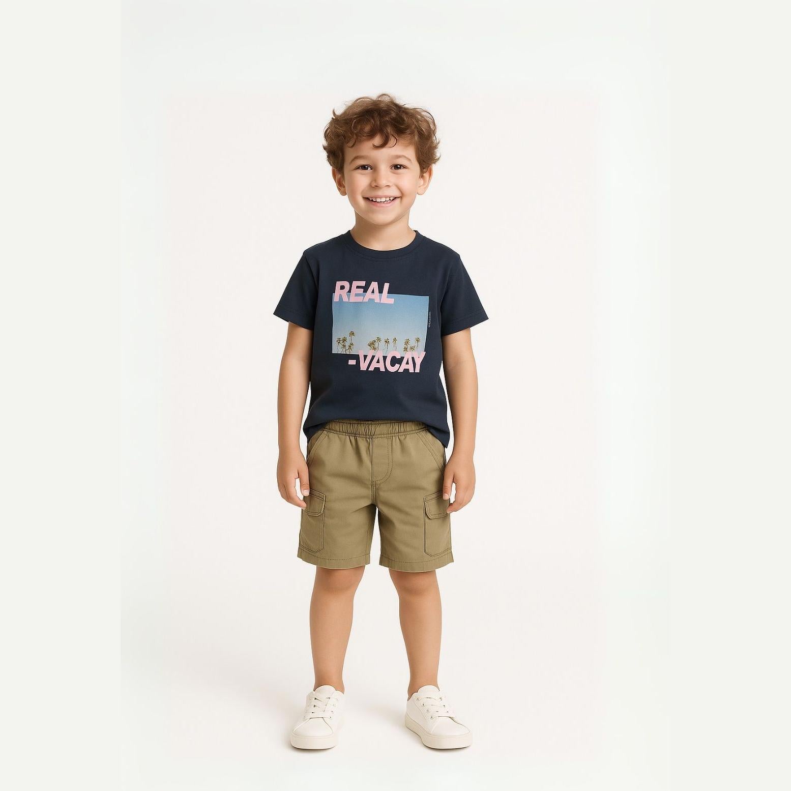 Smiling boy wearing navy blue t-shirt with 'REAL VACATION' graphic and khaki cargo shorts with side pockets, standing against light grey background
