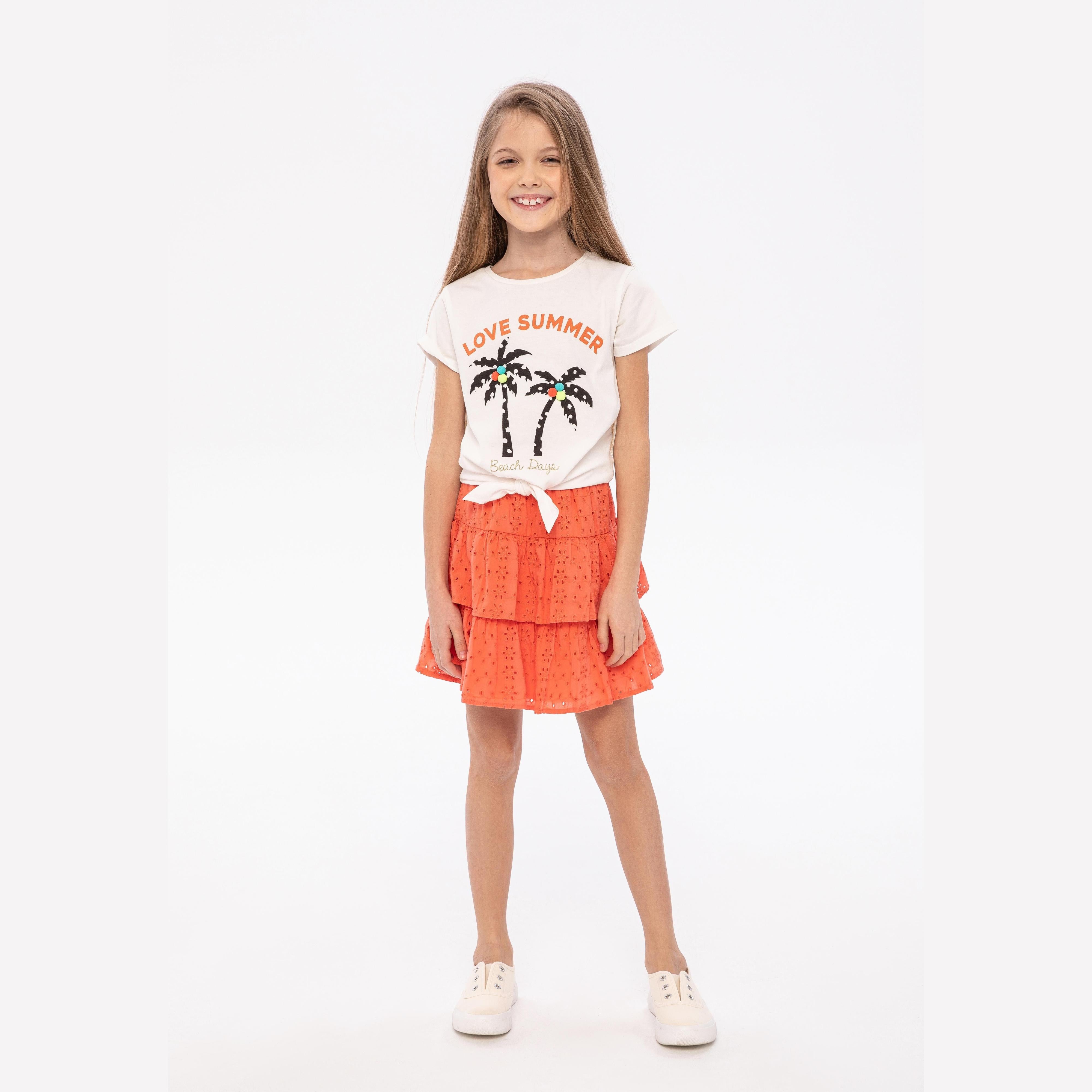 Young girl wearing coral broderie anglaise tiered skirt with white palm tree t-shirt, standing against light background