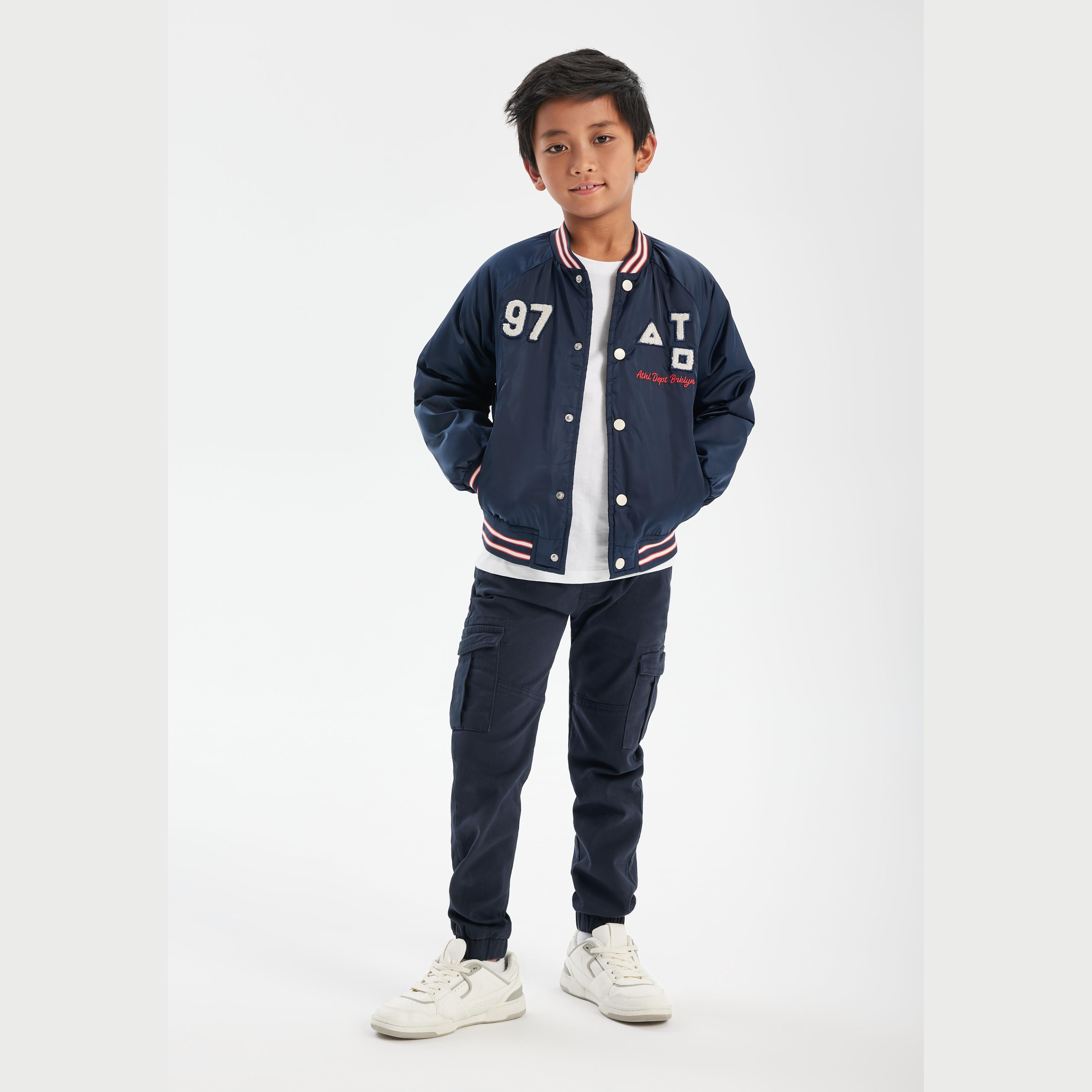 Young boy wearing navy satin bomber jacket with white t-shirt and dark trousers, standing against white background
