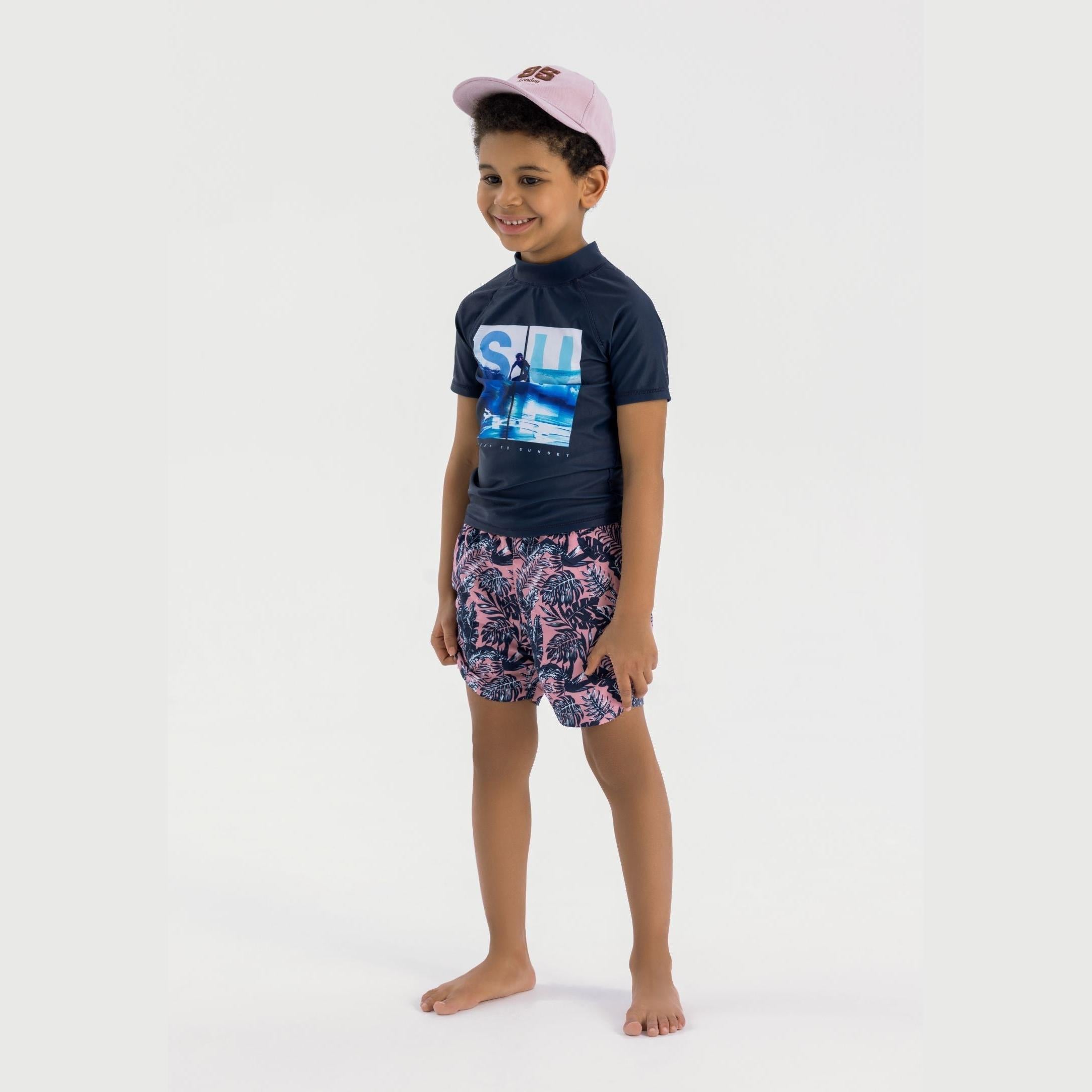 Young boy wearing coral board shorts with tropical leaf print and navy rashguard top, standing barefoot against white background wearing pink cap
