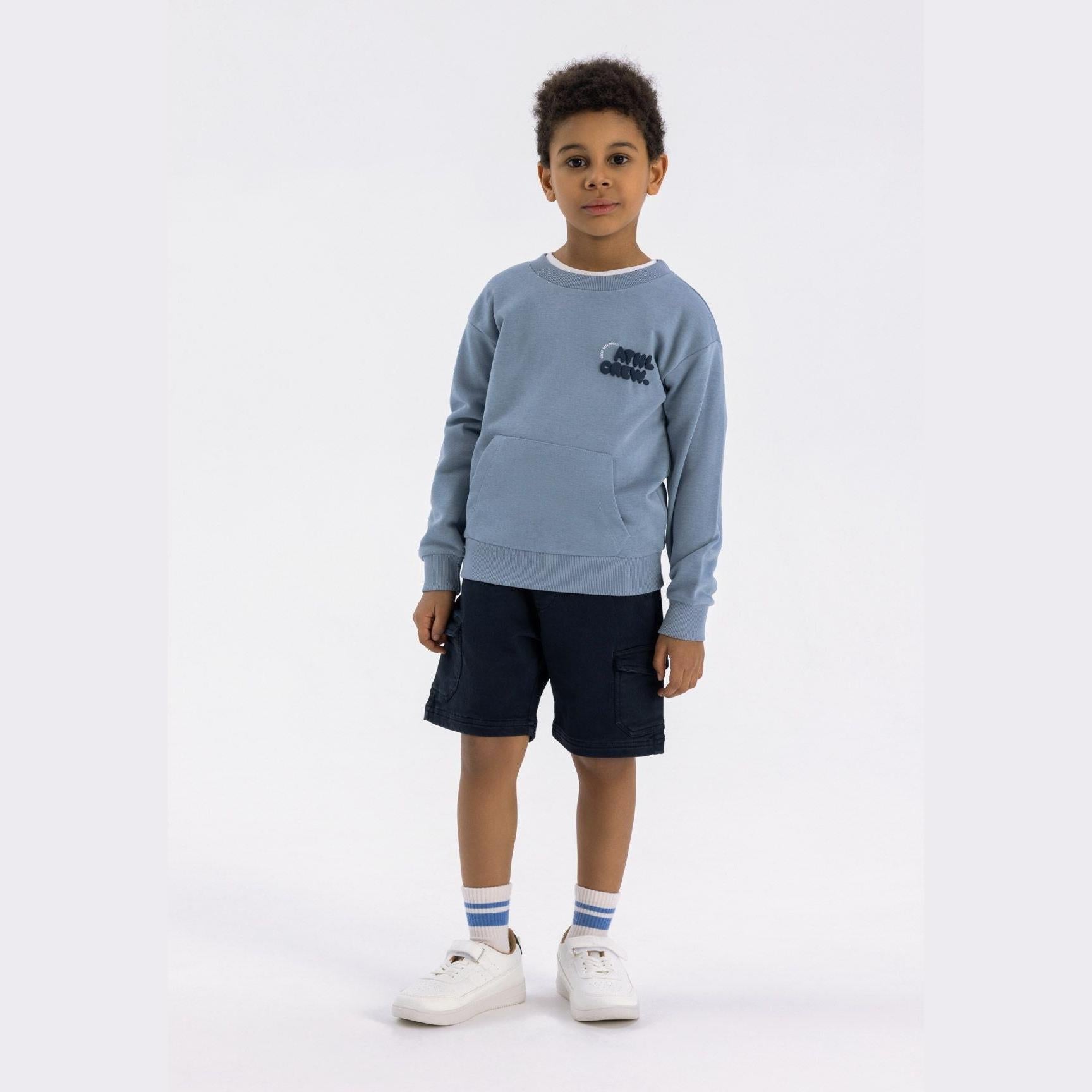 Young boy wearing dusty blue crew neck sweatshirt with kangaroo pocket and dark navy shorts, standing against white background with white sneakers and striped socks