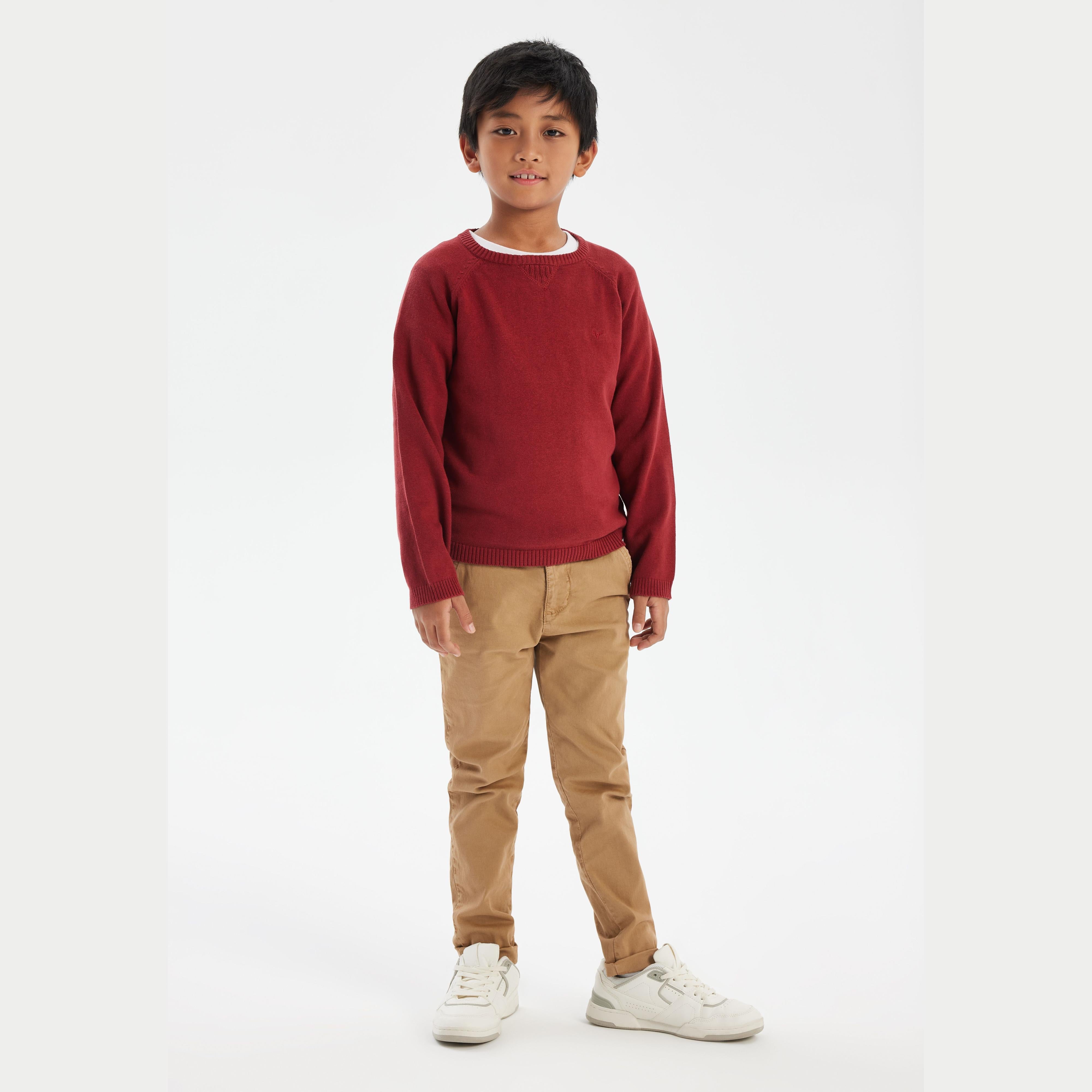 Young boy wearing red knitted crew neck jumper with beige trousers and white trainers, standing against white background